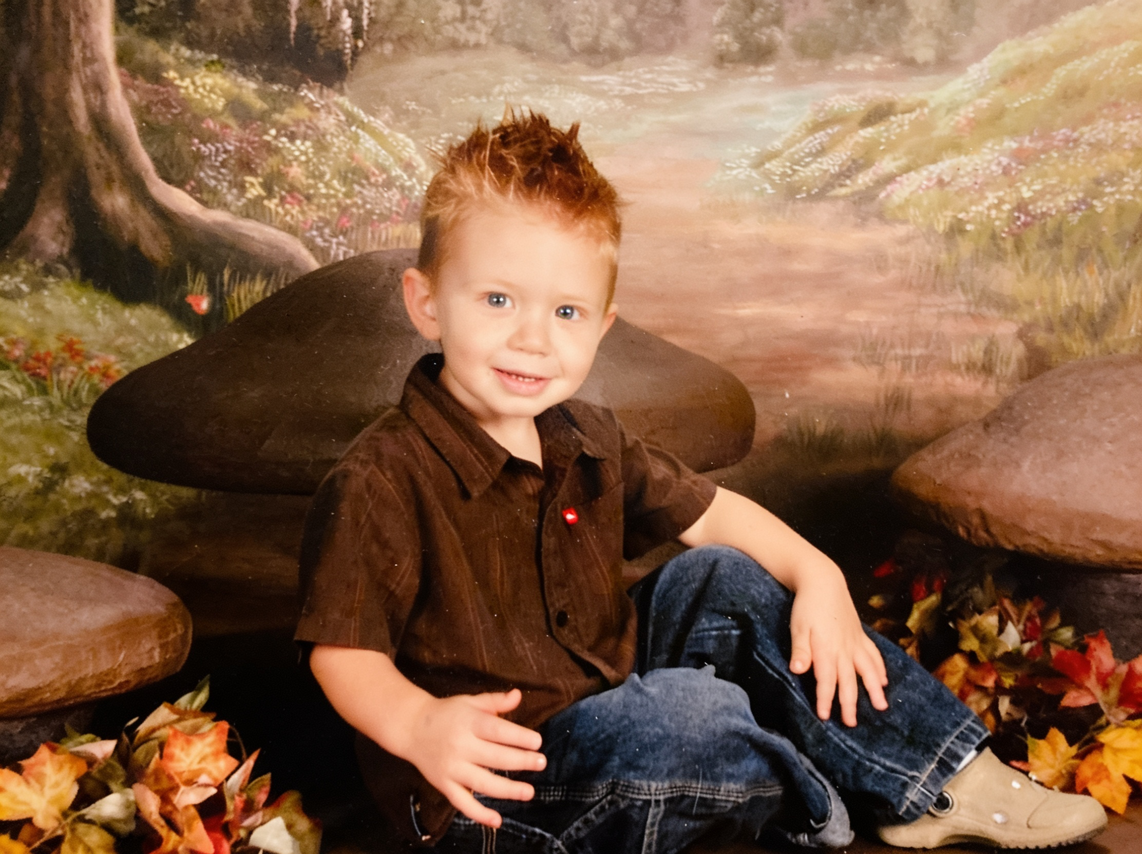 A young boy with red hair and blue eyes smiling, sitting among autumn leaves and rocks, with a painted background of a forest scene.