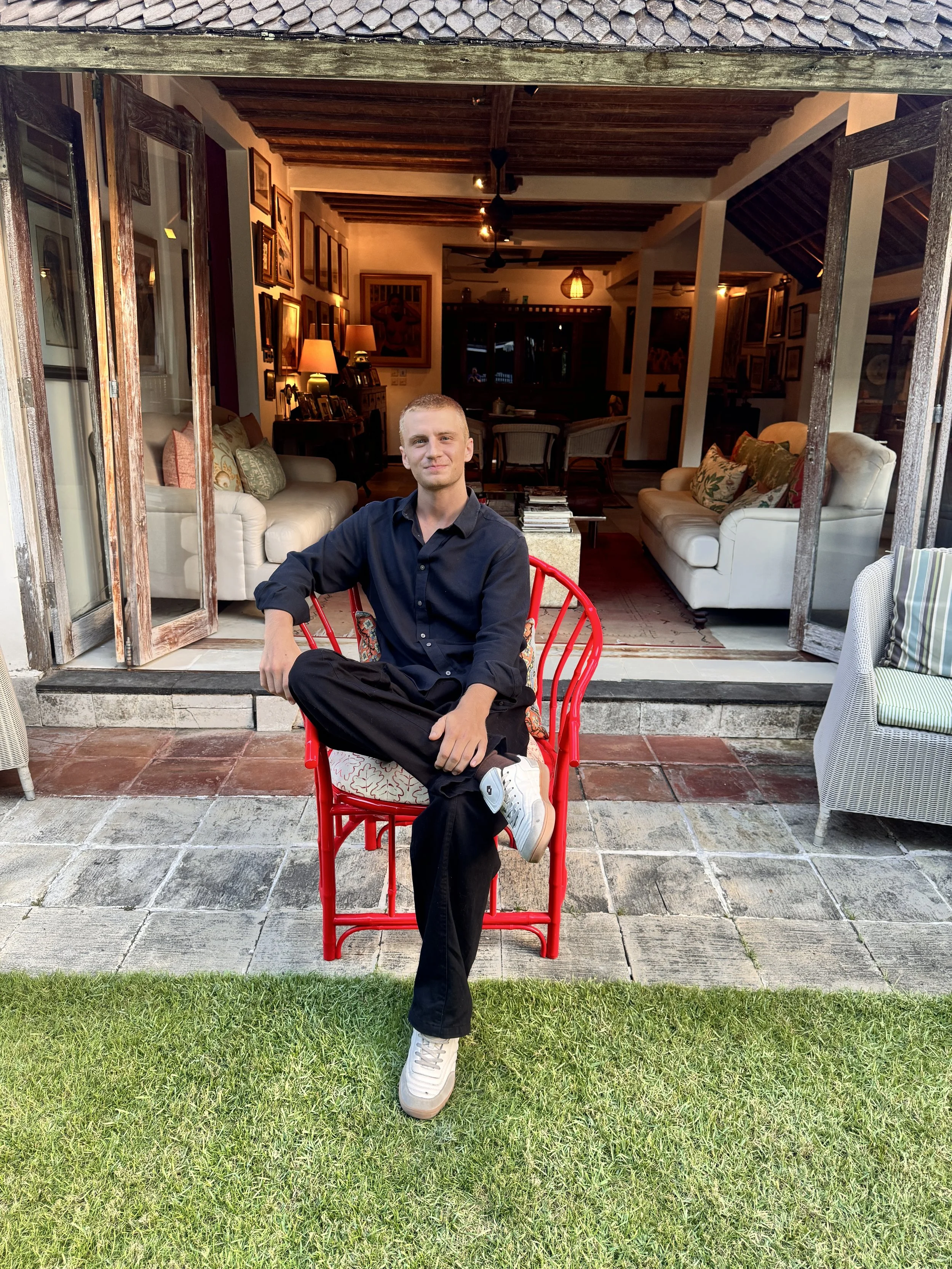 A young man sitting on a red chair outside a house. He is wearing a navy blue shirt, black pants, and white sneakers. The background shows an open sliding door revealing a cozy living room with couches, framed pictures, and lamps.