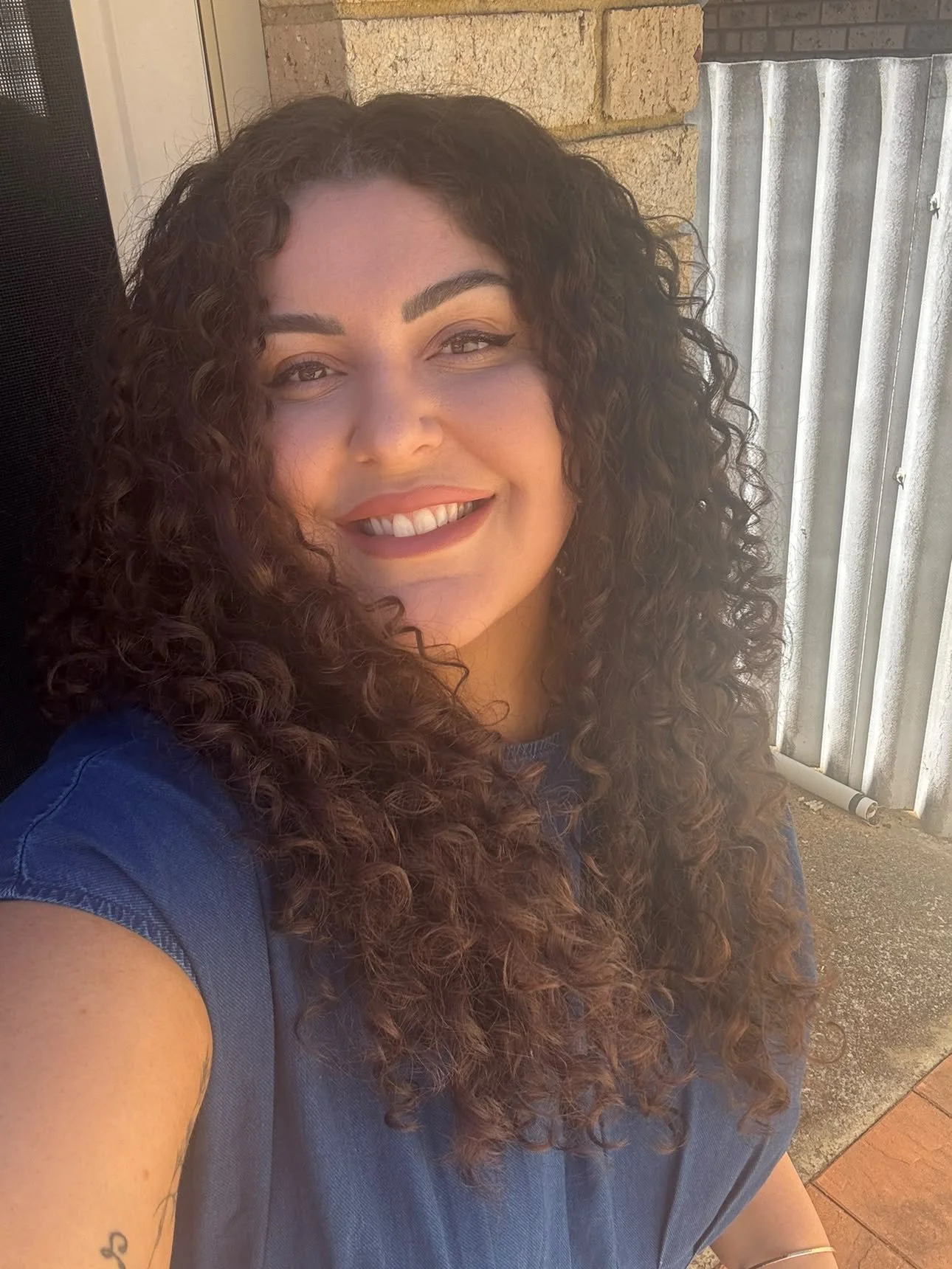 A woman with curly brown hair, wearing a blue top, smiling and taking a selfie outdoors against a background of a brick wall and a corrugated metal door.