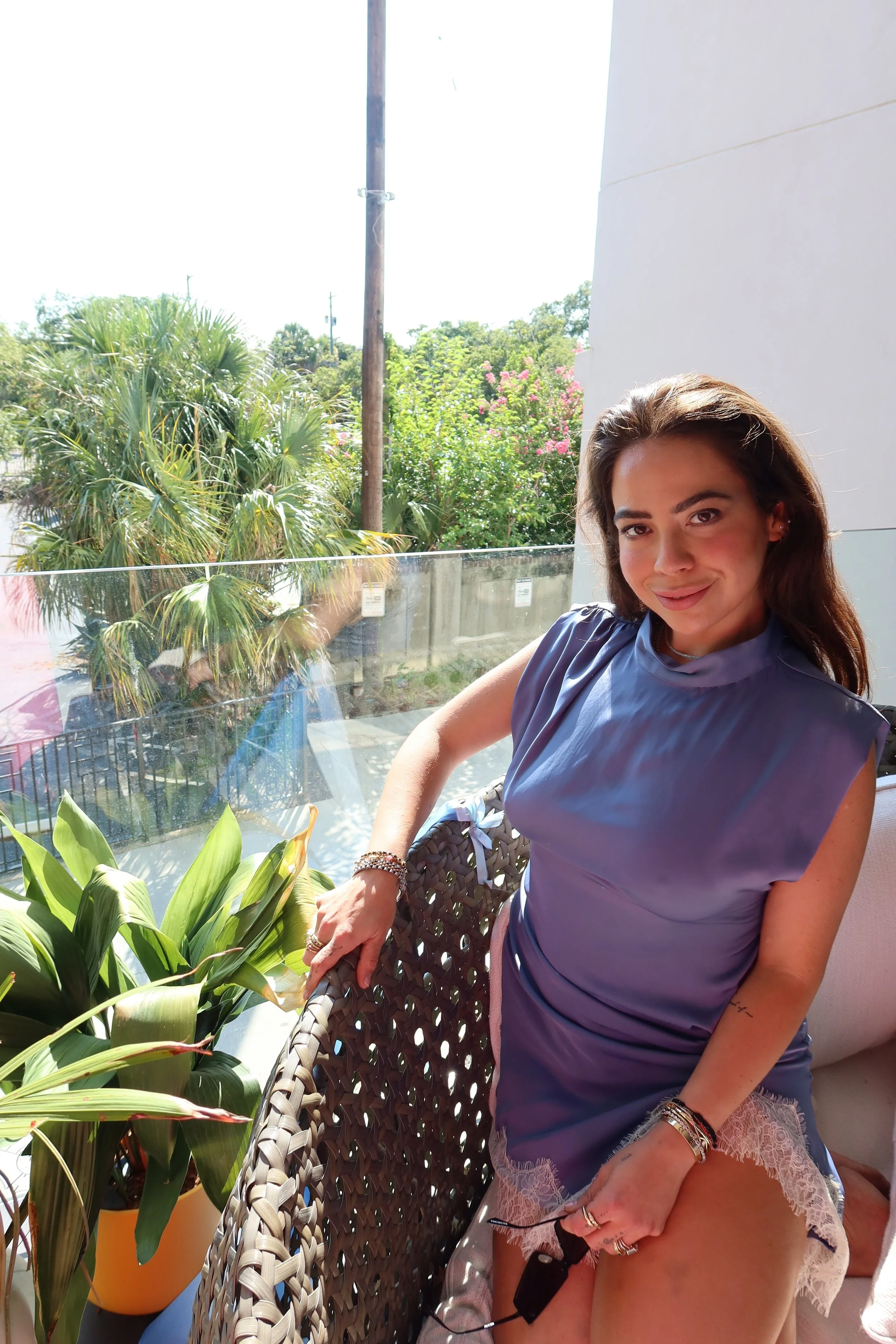 A woman with long brown hair sitting on a balcony with plants, wearing a blue top and pink lace shorts, smiling at the camera.