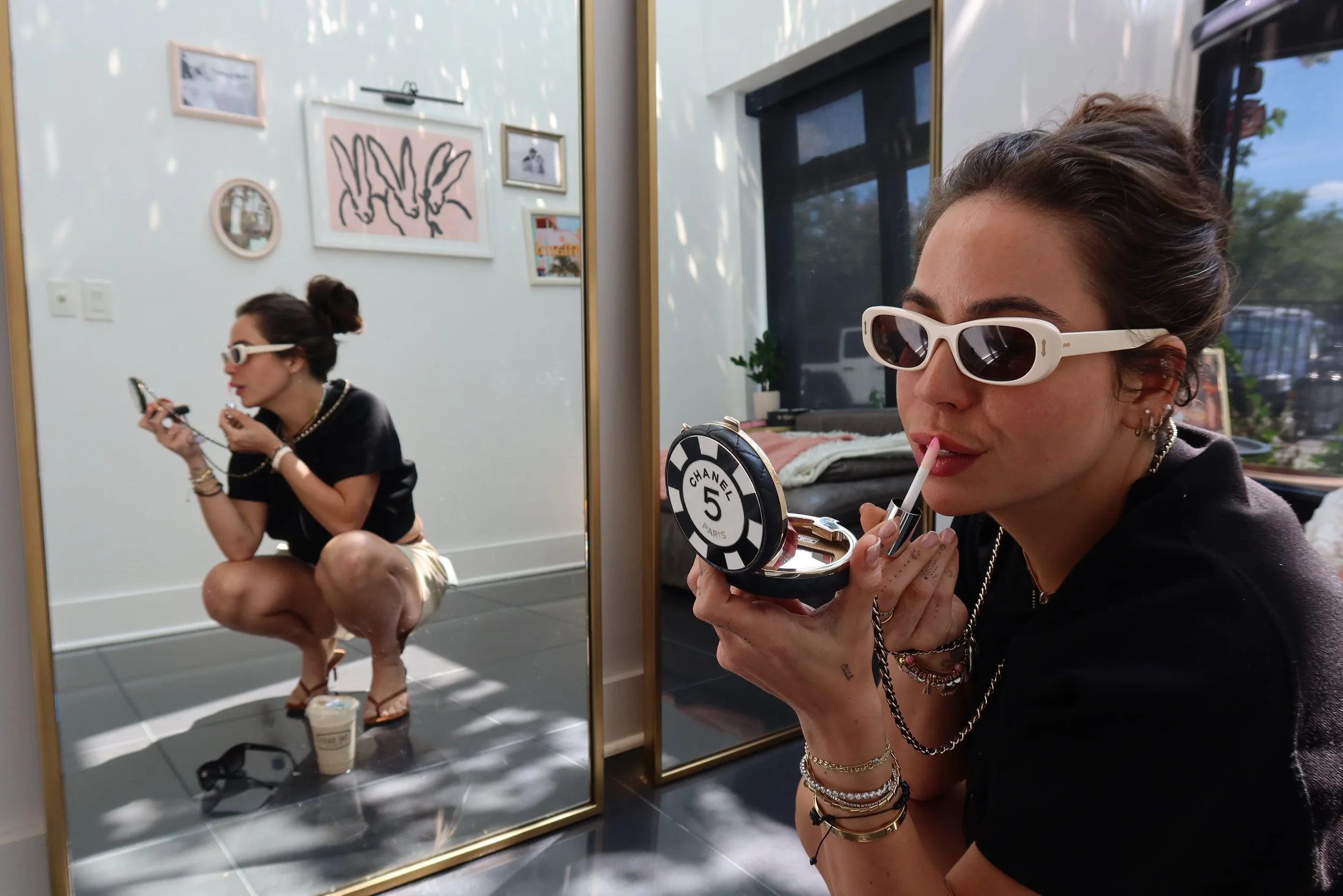 A woman wearing white sunglasses is applying lipstick in front of a large mirror. In the reflection, she is squatting and holding a compact mirror with Chanel branding, and there is a drink cup on the floor. The wall behind her has framed artwork and