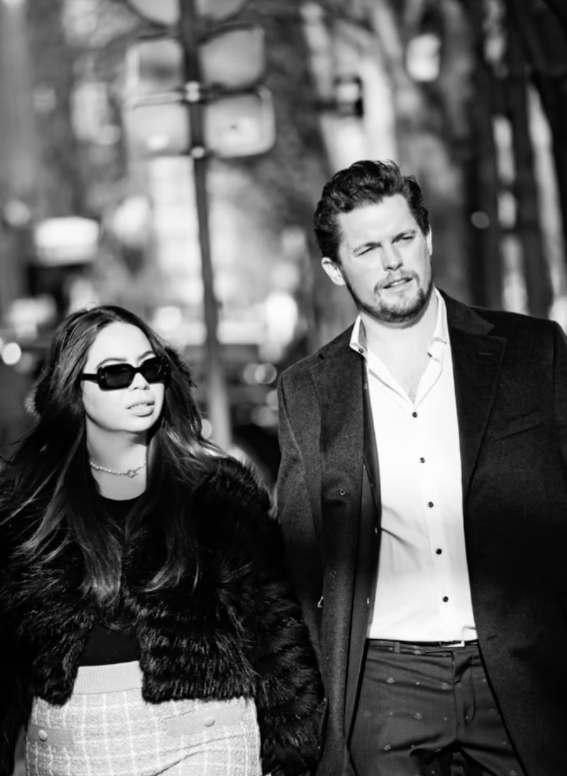 A man and woman walking outdoors in urban setting, black and white photograph. The woman wears sunglasses, a fur coat, and a patterned skirt. The man wears a suit jacket, white shirt, and has a beard and styled hair.