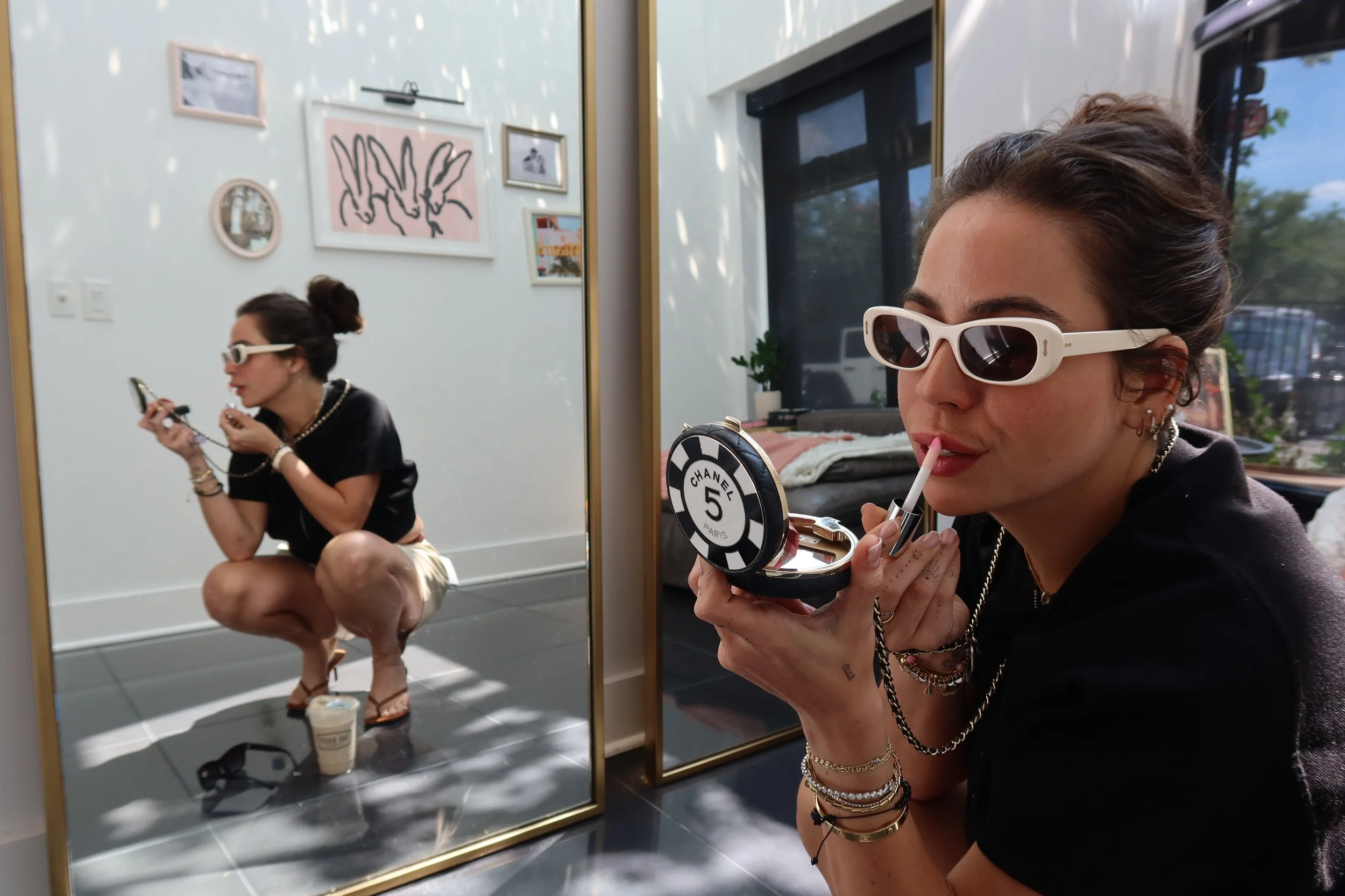 A woman with styled hair and jewelry applying lipstick using a mirror, with her reflection showing her squatting while holding makeup items. She is wearing sunglasses and a black top. The setting is a room with white walls adorned with framed artworks and a window outside with a view of cars and trees.