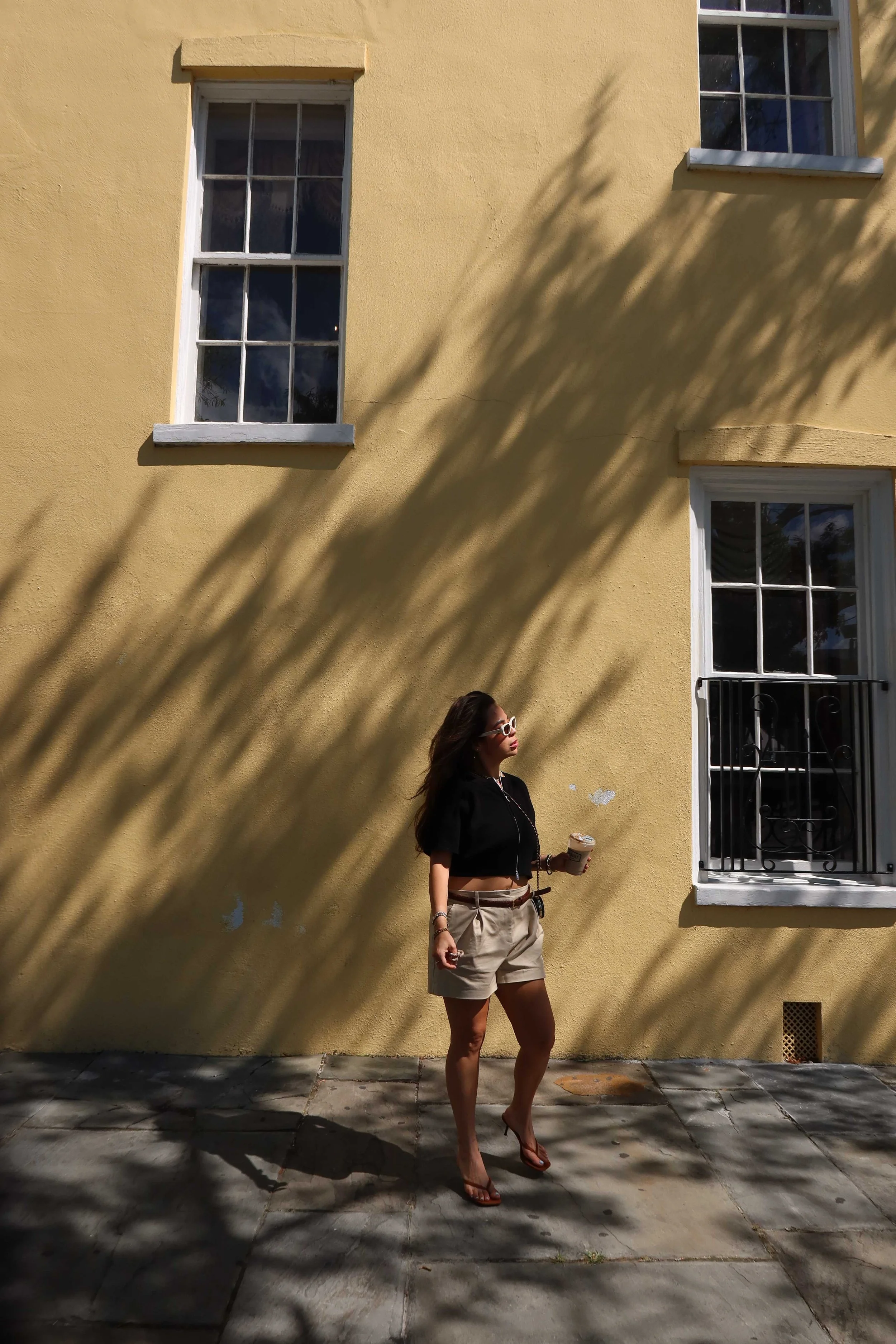 A woman with long dark hair wearing sunglasses, a black cropped jacket, beige shorts, and brown sandals, holding an iced coffee, walking past a yellow building with large windows and casting shadows on the ground.