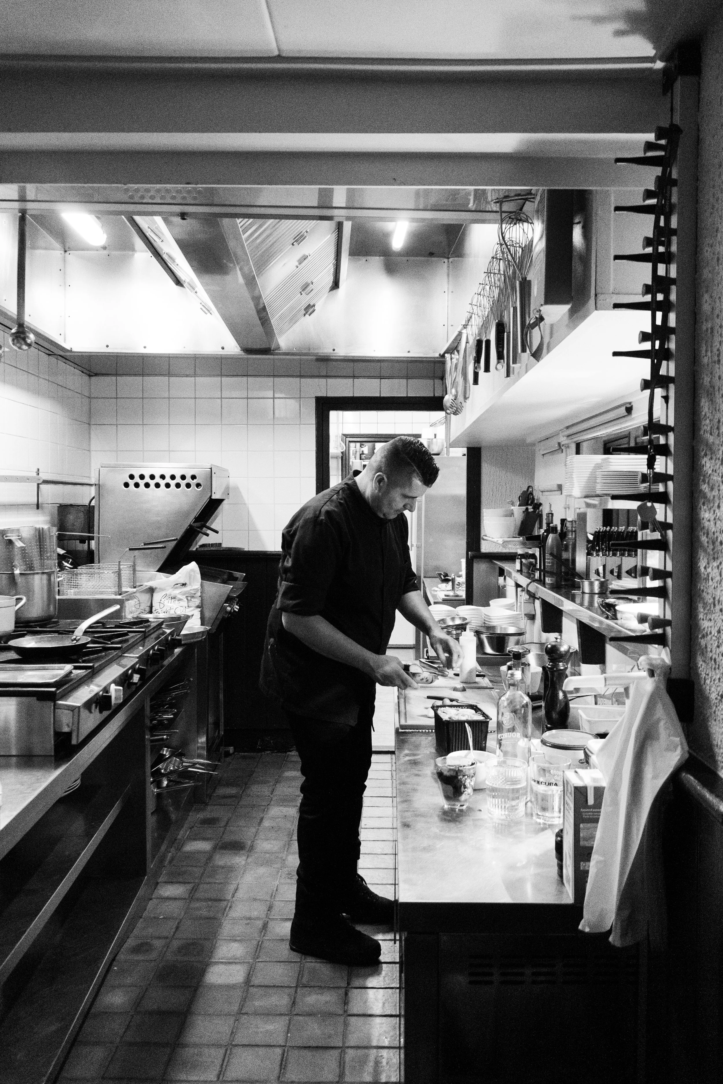 A male chef prepares food in a commercial kitchen, standing at a countertop with various kitchen tools and ingredients. The kitchen has white tiled walls and stainless steel appliances.