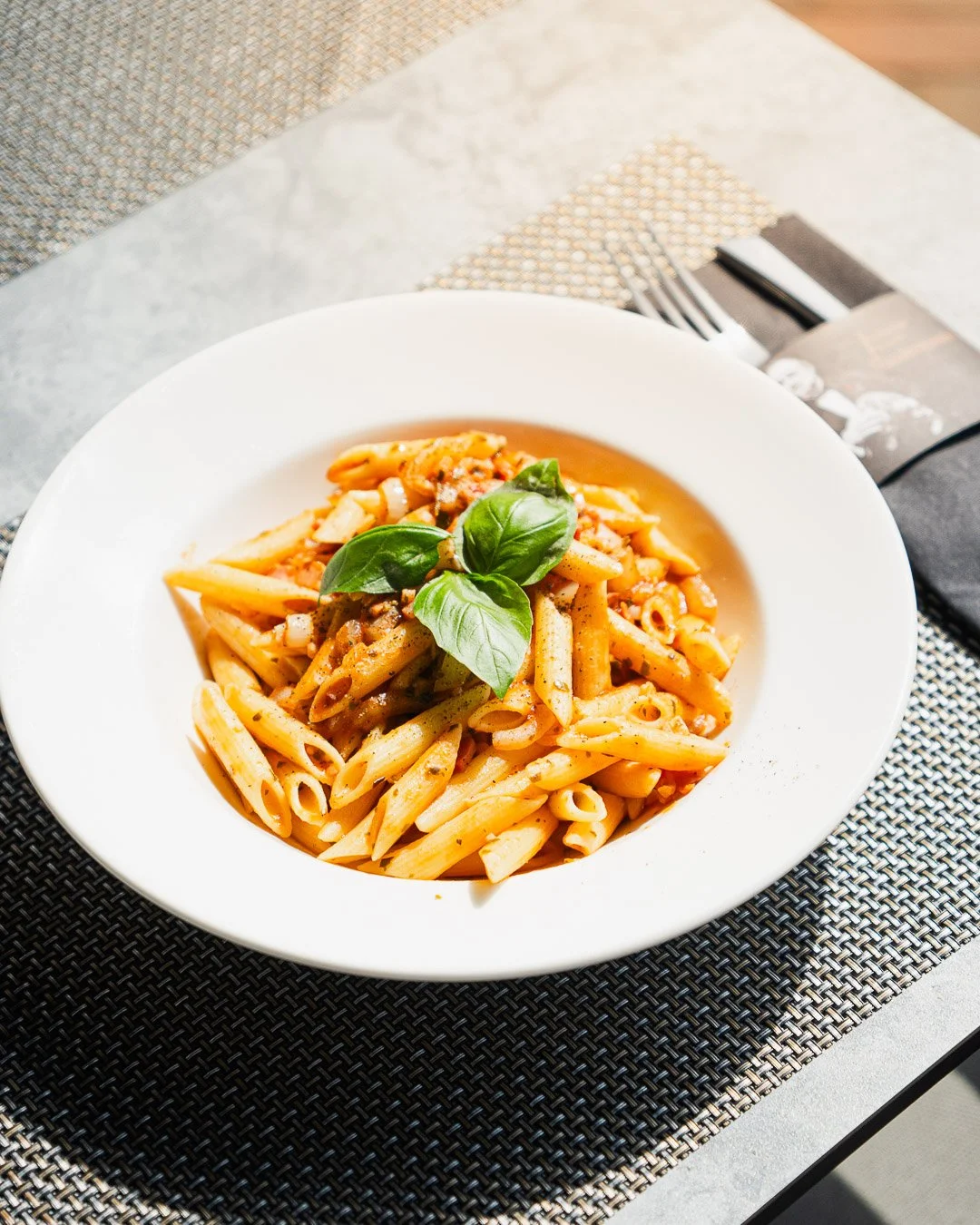 A white bowl of penne pasta with tomato sauce and basil leaves on top, placed on a woven placemat on a table with a fork and napkin nearby.