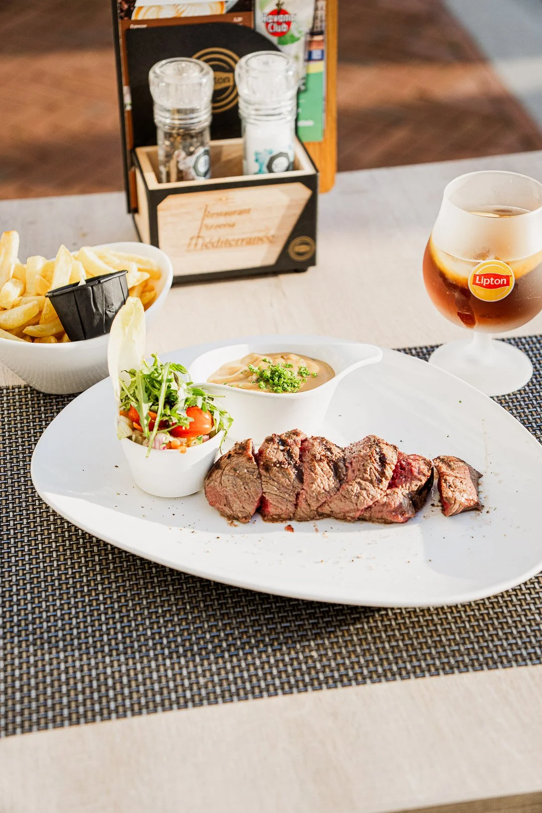 A meal with slices of cooked steak, a small salad with cherry tomatoes and greens, a small dish of sauce or gravy, a bowl of French fries, and a glass of iced tea on a table with a woven placemat.