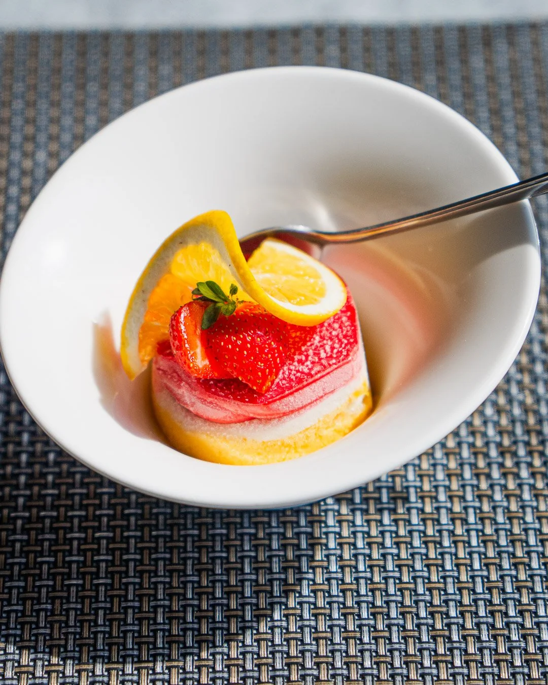 A dessert with a lemon slice, strawberry, and green garnish on top, served in a white bowl with a spoon, on a woven placemat.