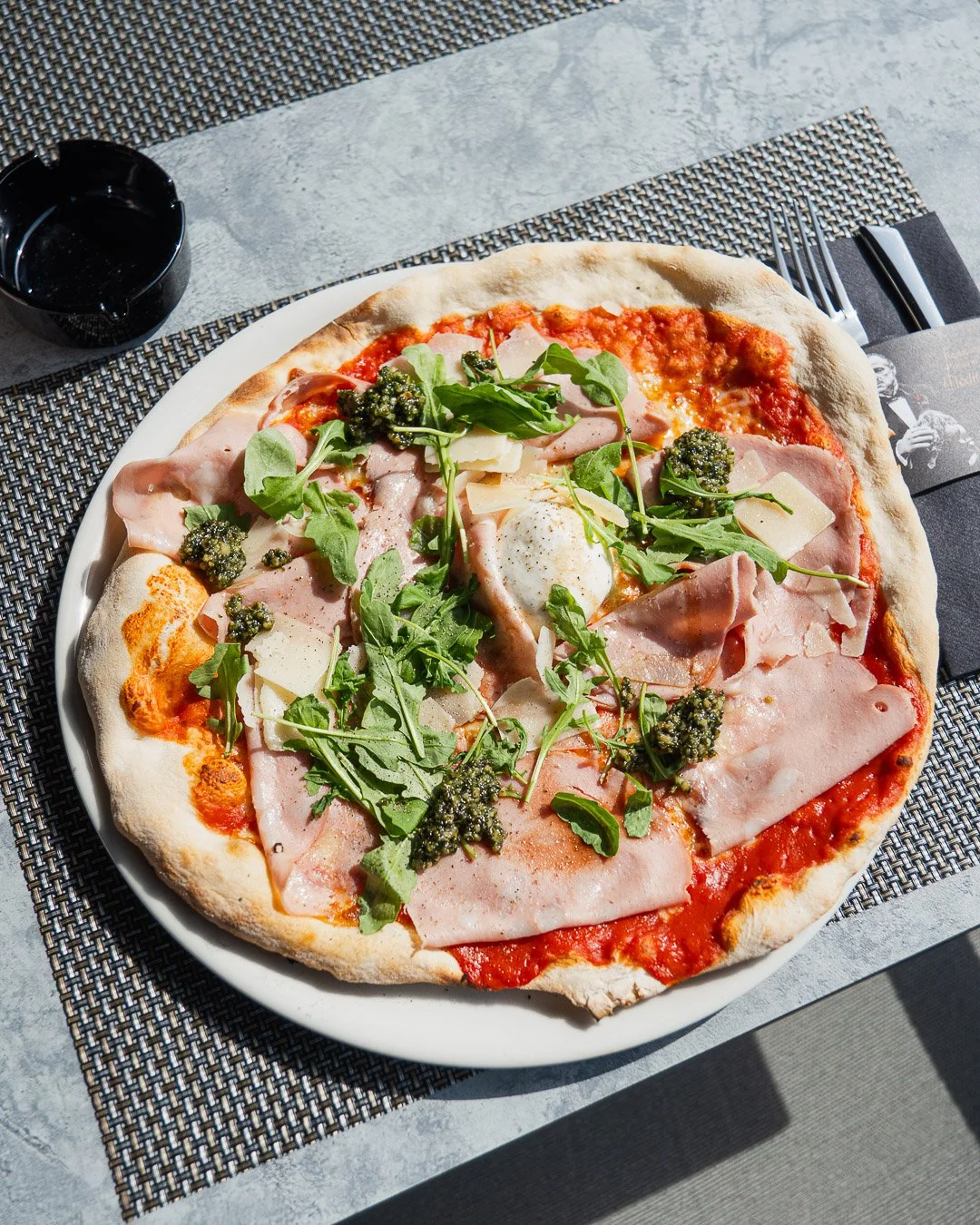 Pizza topped with ham, arugula, cheese, and basil pesto on a white plate, with a small black dish of balsamic vinegar nearby.