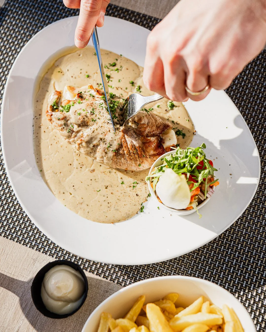 A plate with a roasted meat dish topped with creamy white sauce being cut with a fork and knife, accompanied by a small bowl of salad, a side of fries, and a small container of mayonnaise.