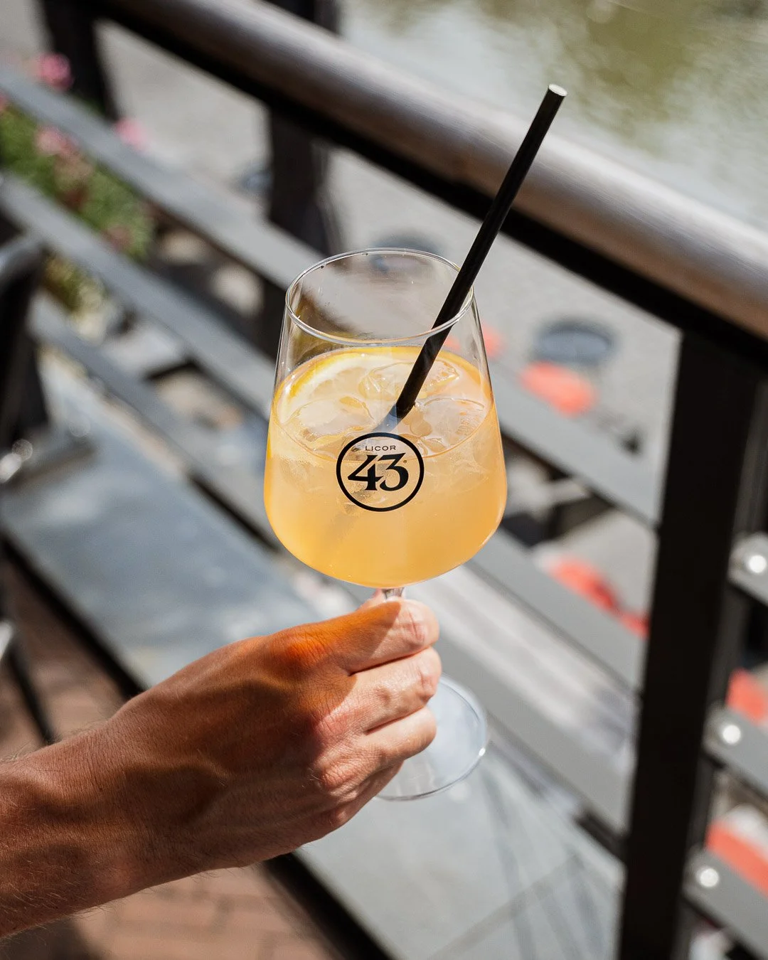 A hand holding a glass of orange-colored cocktail with ice and a black straw, with 'LICOR 43' logo on the glass, against a background of a wooden railing and sidewalk.