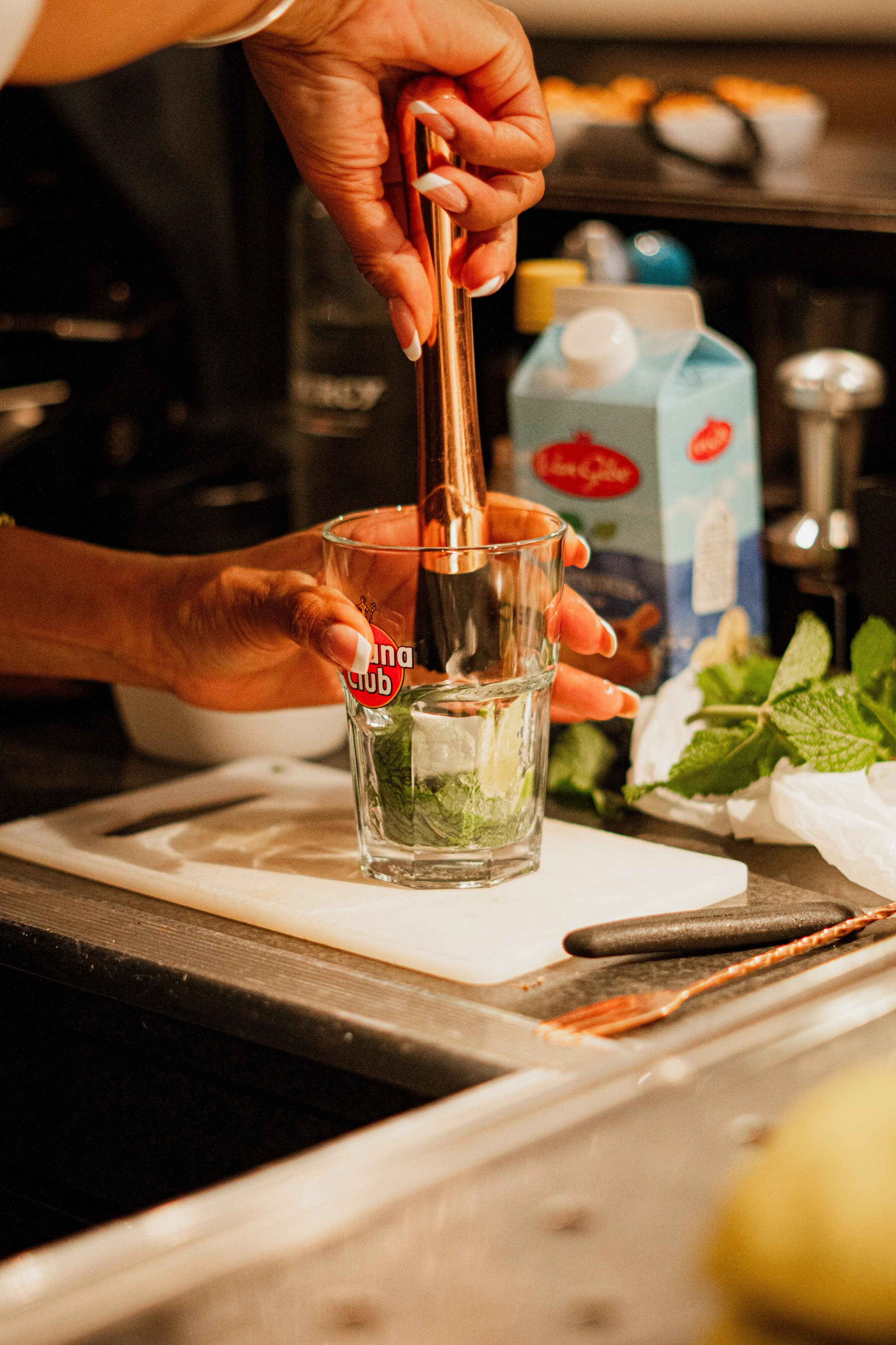 Person using a copper bar to muddle fresh mint leaves in a glass for making a beverage. The scene is in a kitchen with various ingredients like a carton of cream and a bowl of lettuce on the counter.