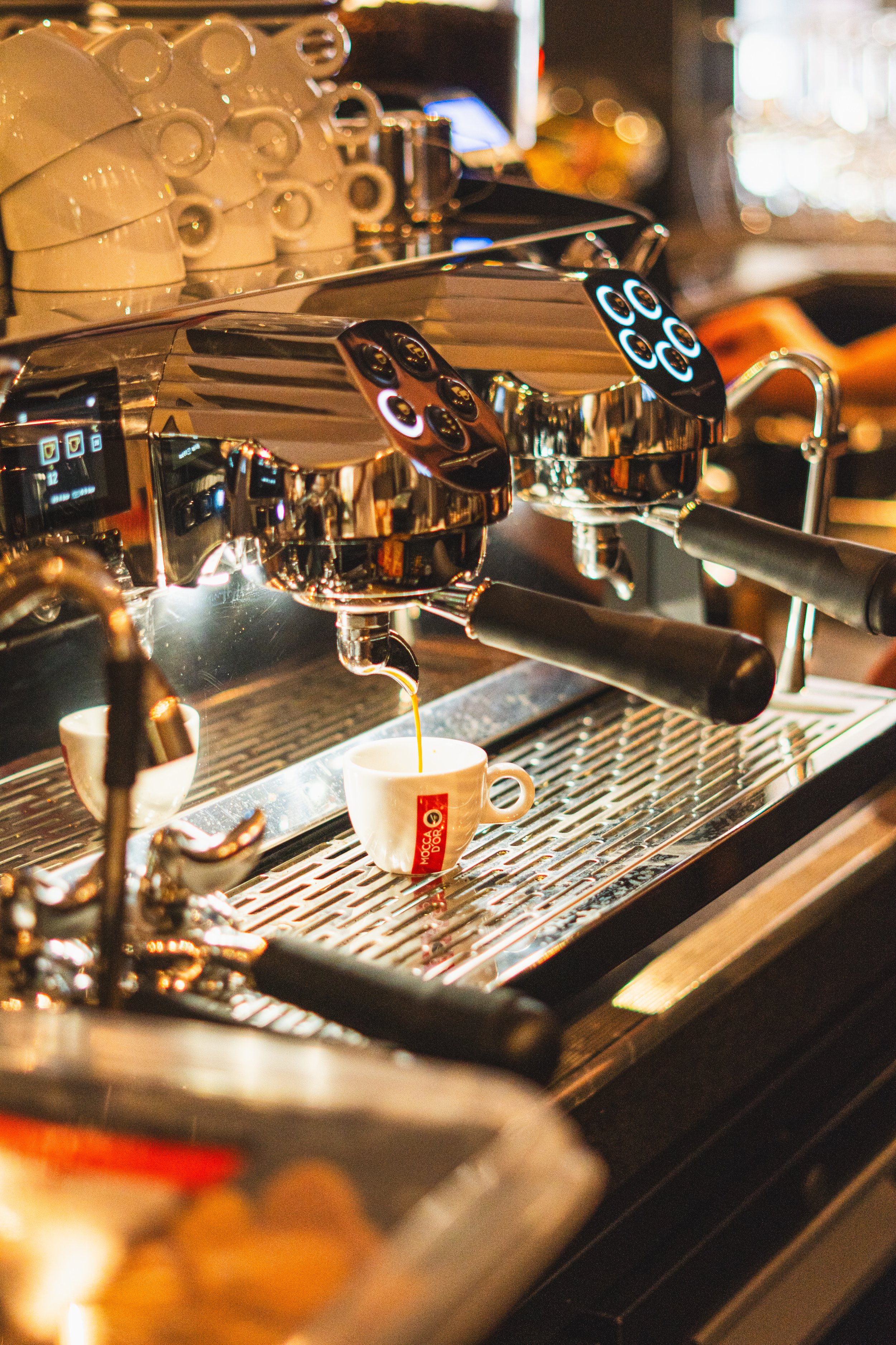 A shiny espresso machine brewing coffee into a white cup with the logo 'MoccaD'Oro' on it, with cups stacked in the background.