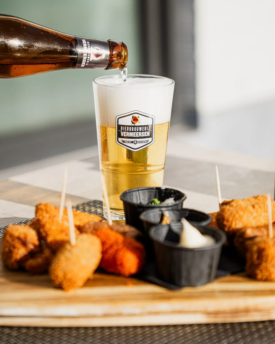 A glass of beer with frothy head being poured from a bottle, served with fried appetizers and dipping sauces on a wooden platter.