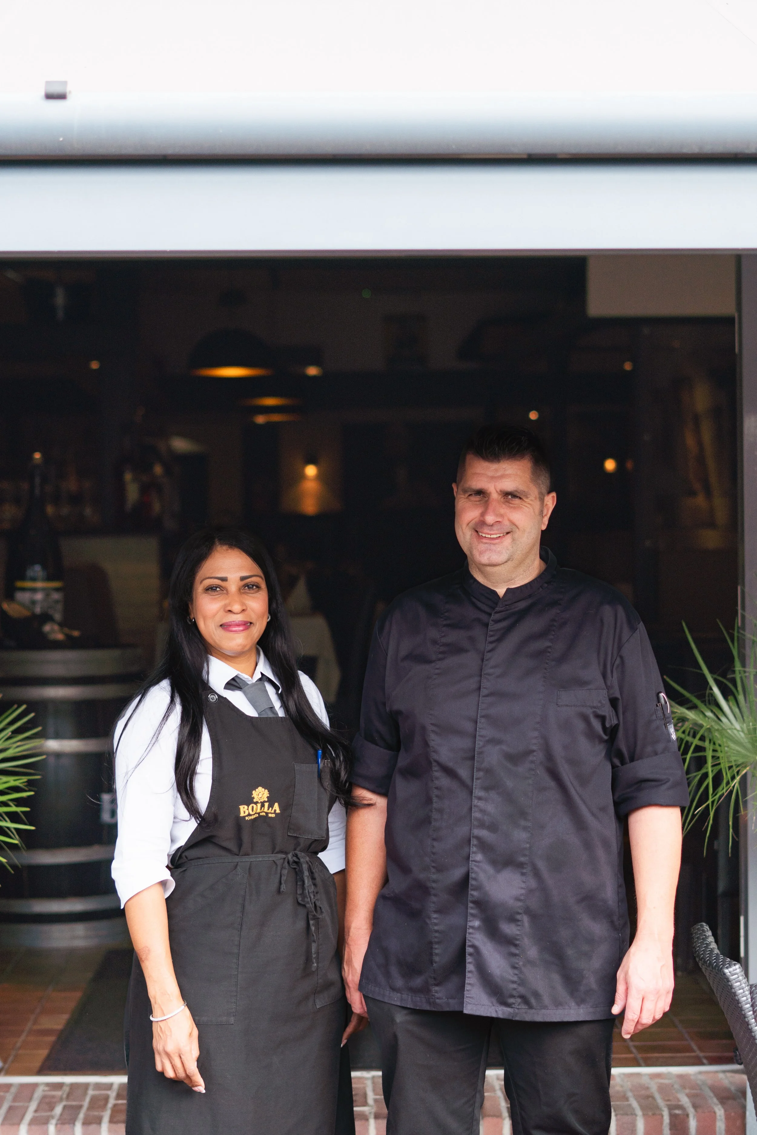 Two restaurant staff members are standing outdoors in front of a restaurant entrance, smiling at the camera. The woman on the left is wearing a white shirt and a black apron with the word "BOLLA" embroidered on it, while the man on the right is wearing a black chef's jacket.