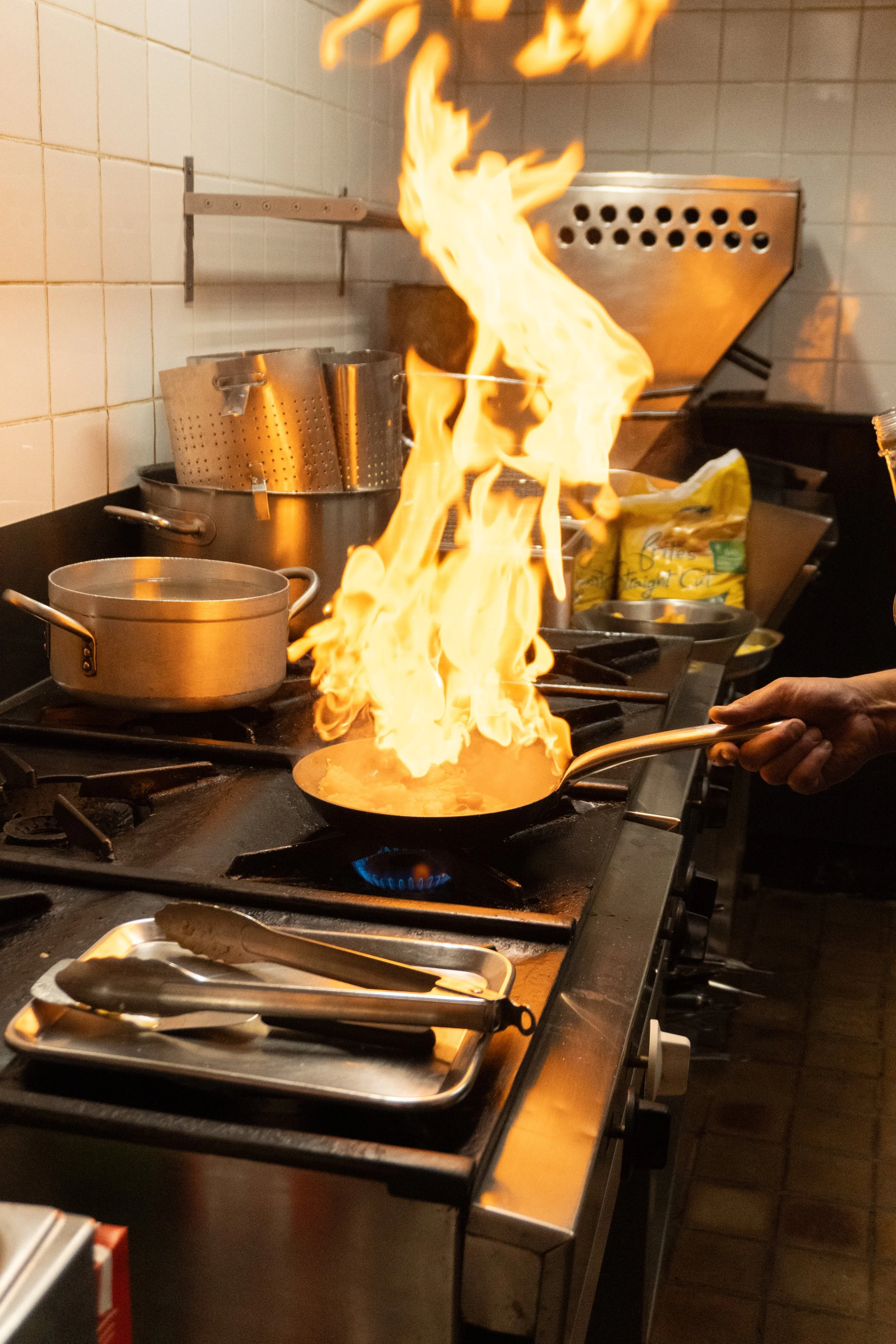 A kitchen stove with a flaming pan and other cookware, including a large pot, with a tiled backsplash.