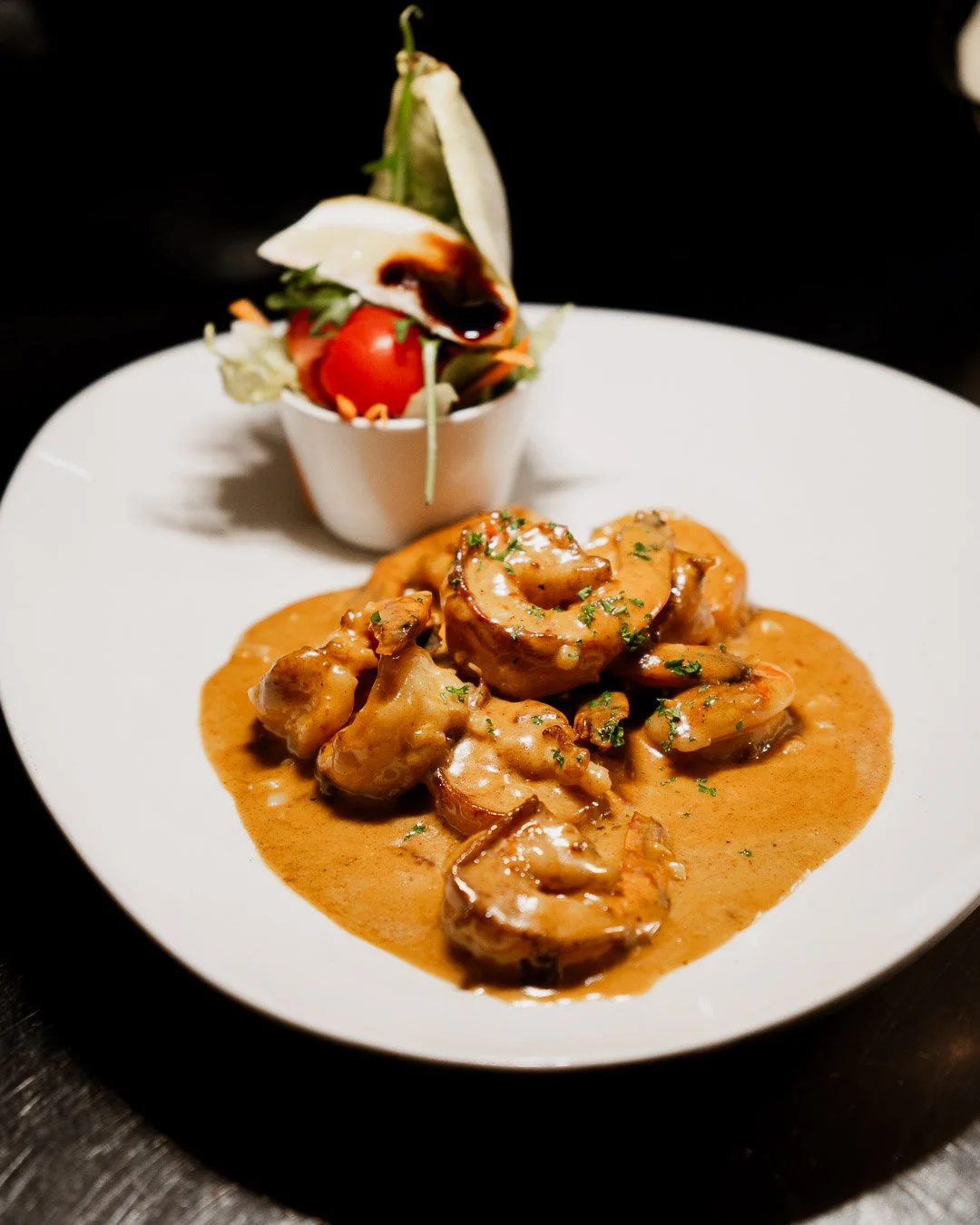 Shrimp and chicken in a creamy sauce served on a white plate, with a side salad containing cherry tomato, lettuce, cucumber, and shredded carrots in a small cup.