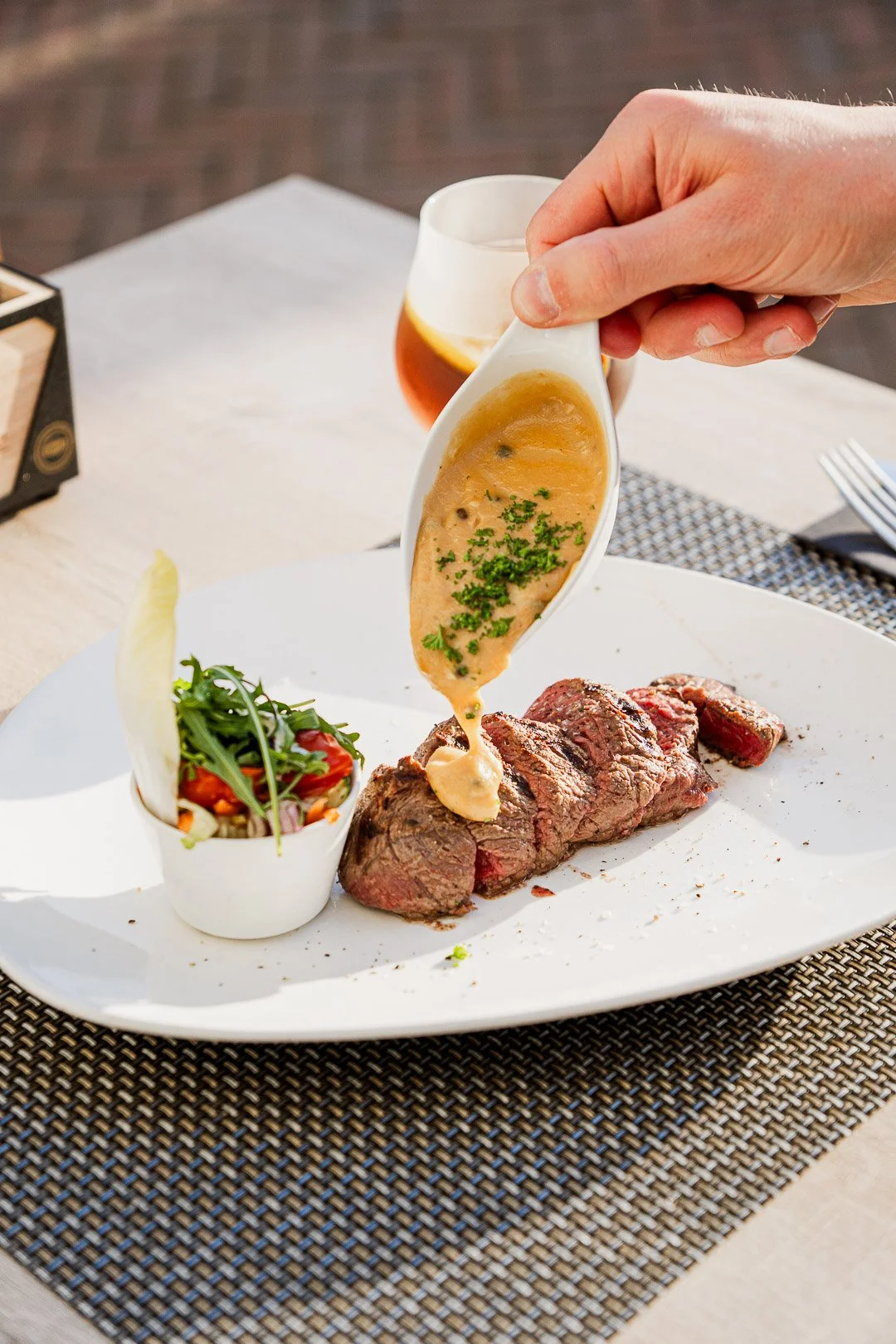 A plate with slices of cooked steak, topped with mustard sauce, alongside a small salad with greens and cherry tomatoes, on a woven placemat.