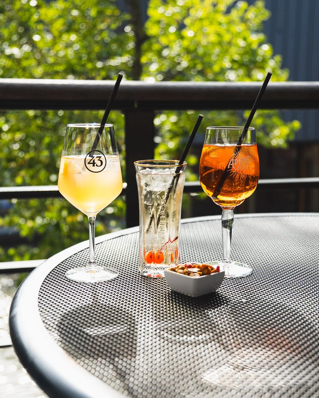 Three drinks on a mesh outdoor table with a small bowl of mixed nuts.