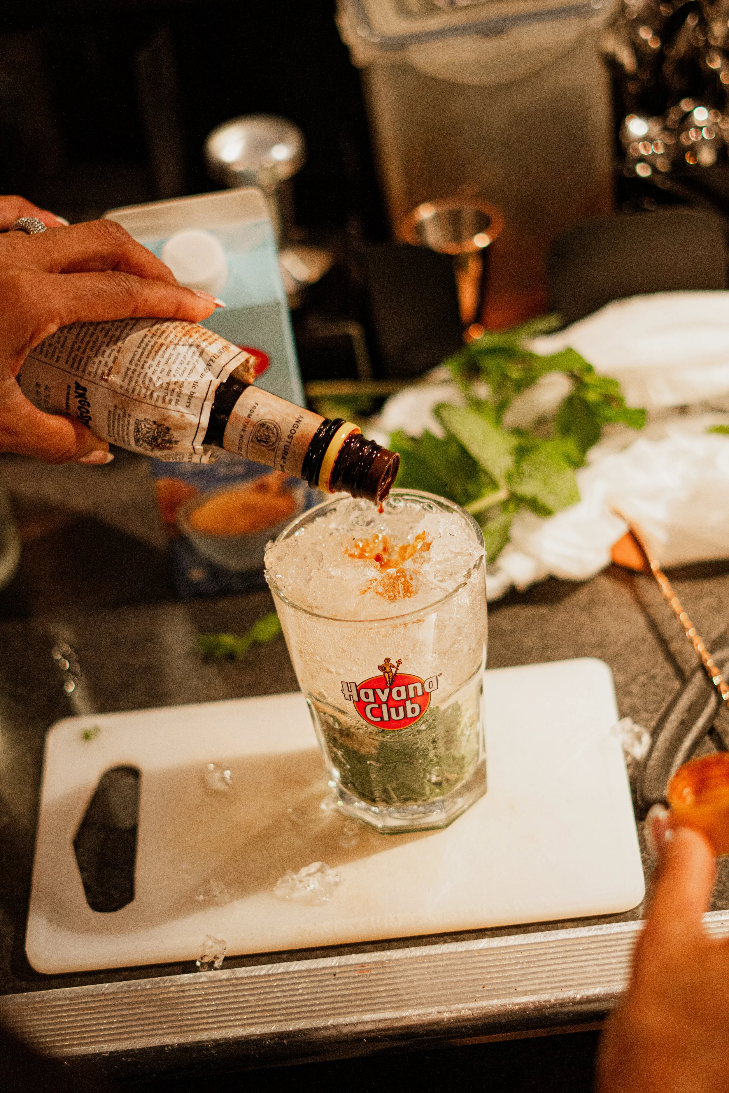 A hand pouring a liquid from a bottle into a glass with ice and a Havana Club logo on it. The background shows a countertop with some green herbs, a white cutting board, and blurred kitchen items.