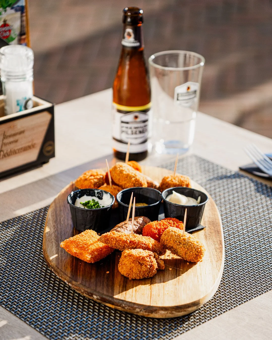 A wooden platter with fried finger foods and dipping sauces on a dining table, with a beer bottle, glass of water, and condiments in the background.