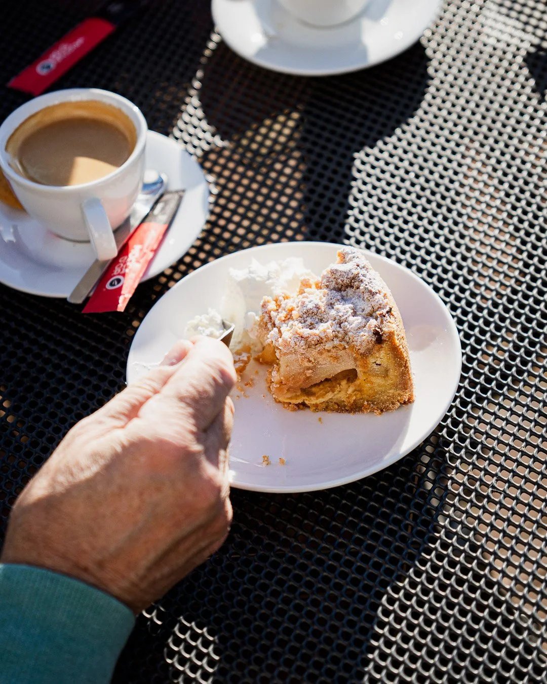 A person's hand holding a spoon over a plate with a slice of apple pie topped with powdered sugar, accompanied by a scoop of vanilla ice cream. On the table, there is a cup of coffee, and a sugar packet and spoon are visible. The table surface is bla