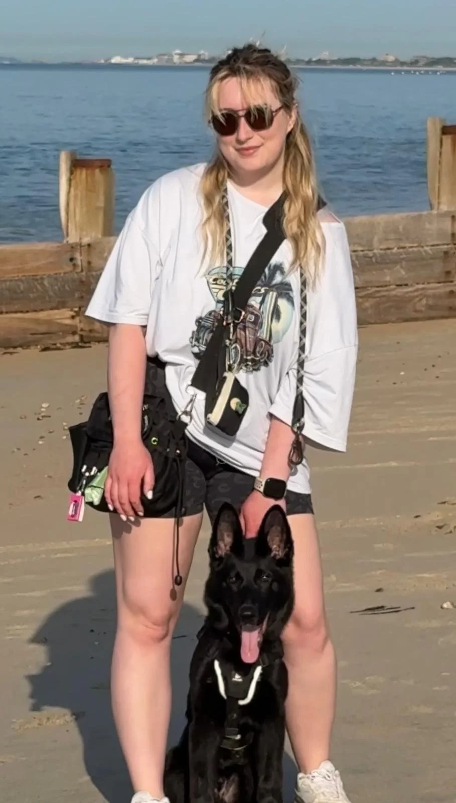 A woman with blonde hair, wearing sunglasses, a white graphic T-shirt, and black shorts, standing on a sandy beach holding a black dog by the leash. The background shows water and a wooden barrier, with a distant shoreline.