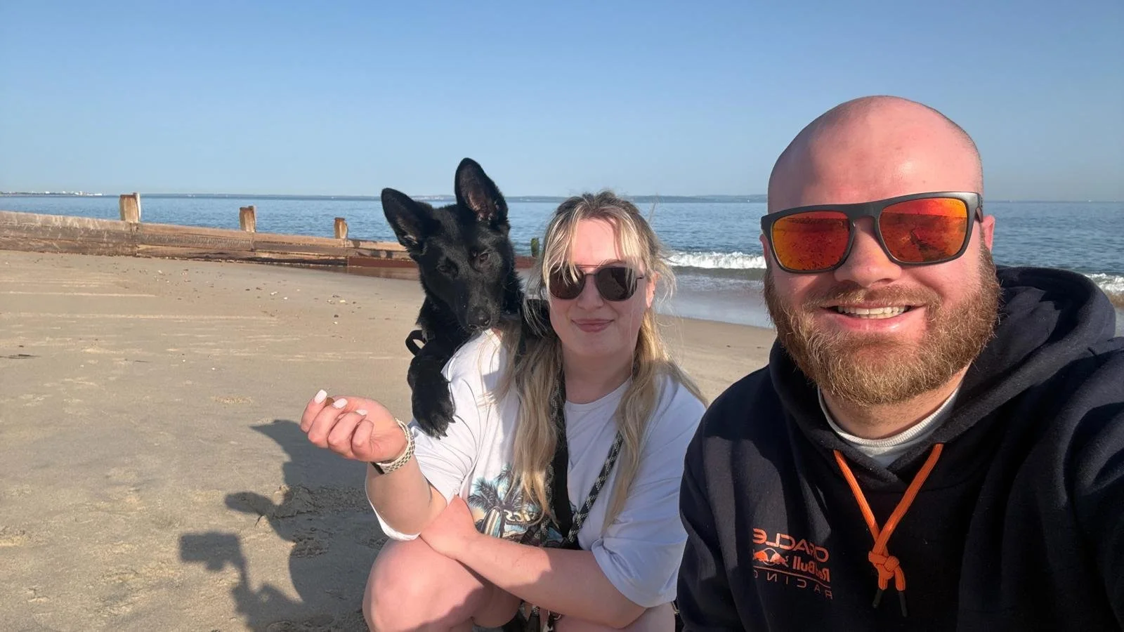 A smiling man with a beard and sunglasses taking a selfie on the beach, holding a black dog, with a woman wearing sunglasses in the background. The beach has sand and a wooden barrier, with the ocean and blue sky in the background.