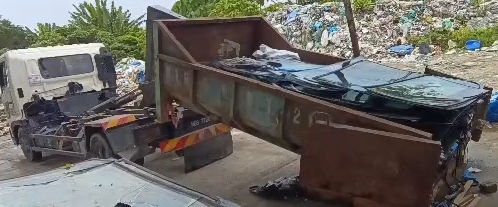 A damaged dump truck in a junkyard with debris and other vehicles around.