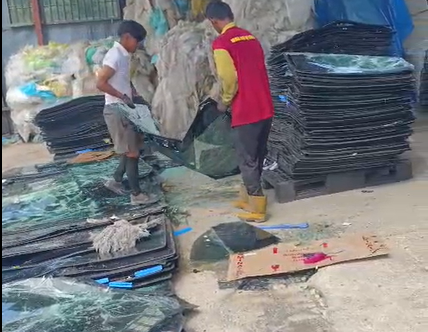 Two workers handling broken glass and plastic sheets near stacked black plastic containers outdoors.