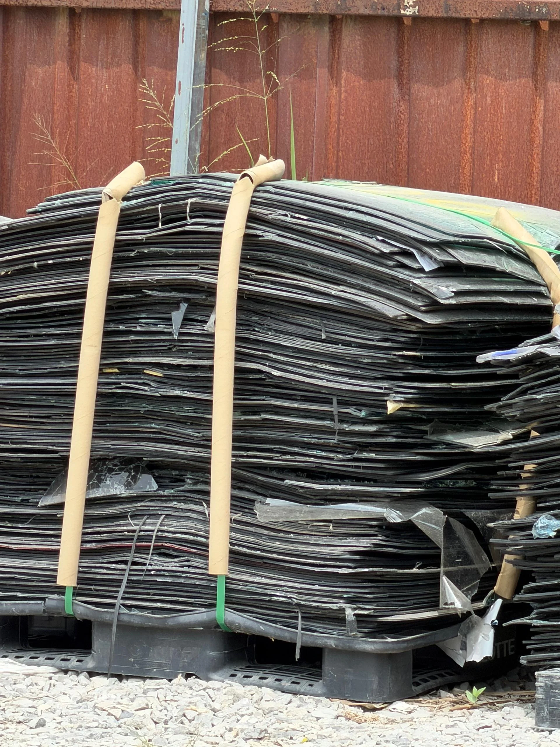 A large bundle of black cables tied with beige straps, resting on a black plastic pallet outside against a rusty metal fence, with some grass and small plants nearby.
