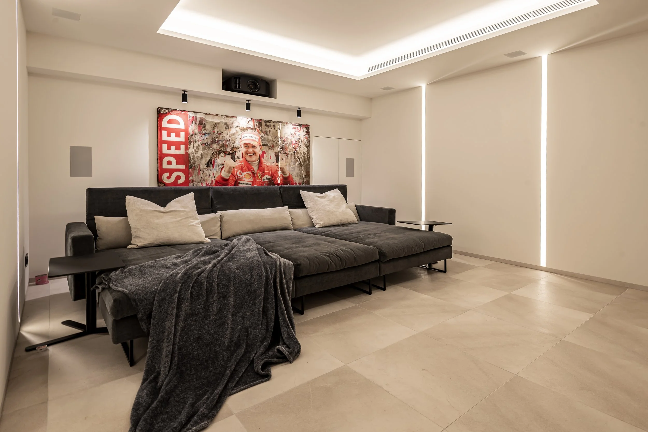 Sala de estar moderna con sofá gris y almohadas blancas, una manta gris, mesas pequeñas y una pared decorada con una imagen de un piloto de Fórmula 1 sonriendo con un fondo de graffiti y la palabra 'SPEED' en letras rojas, iluminación suave y paredes