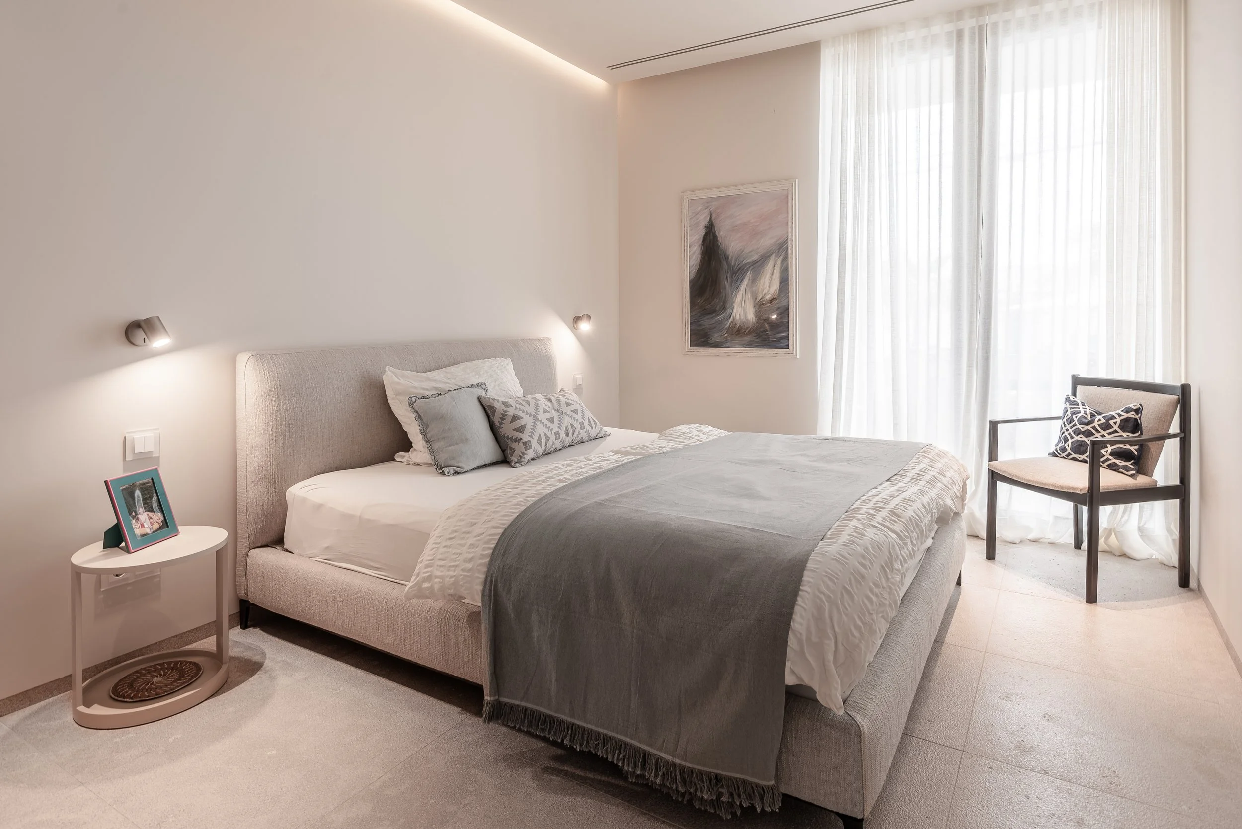Habitación moderna con cama grande, almohadas decorativas, mesita de noche con marco de fotos, cortinas blancas y pared decorativa con pintura abstracta.