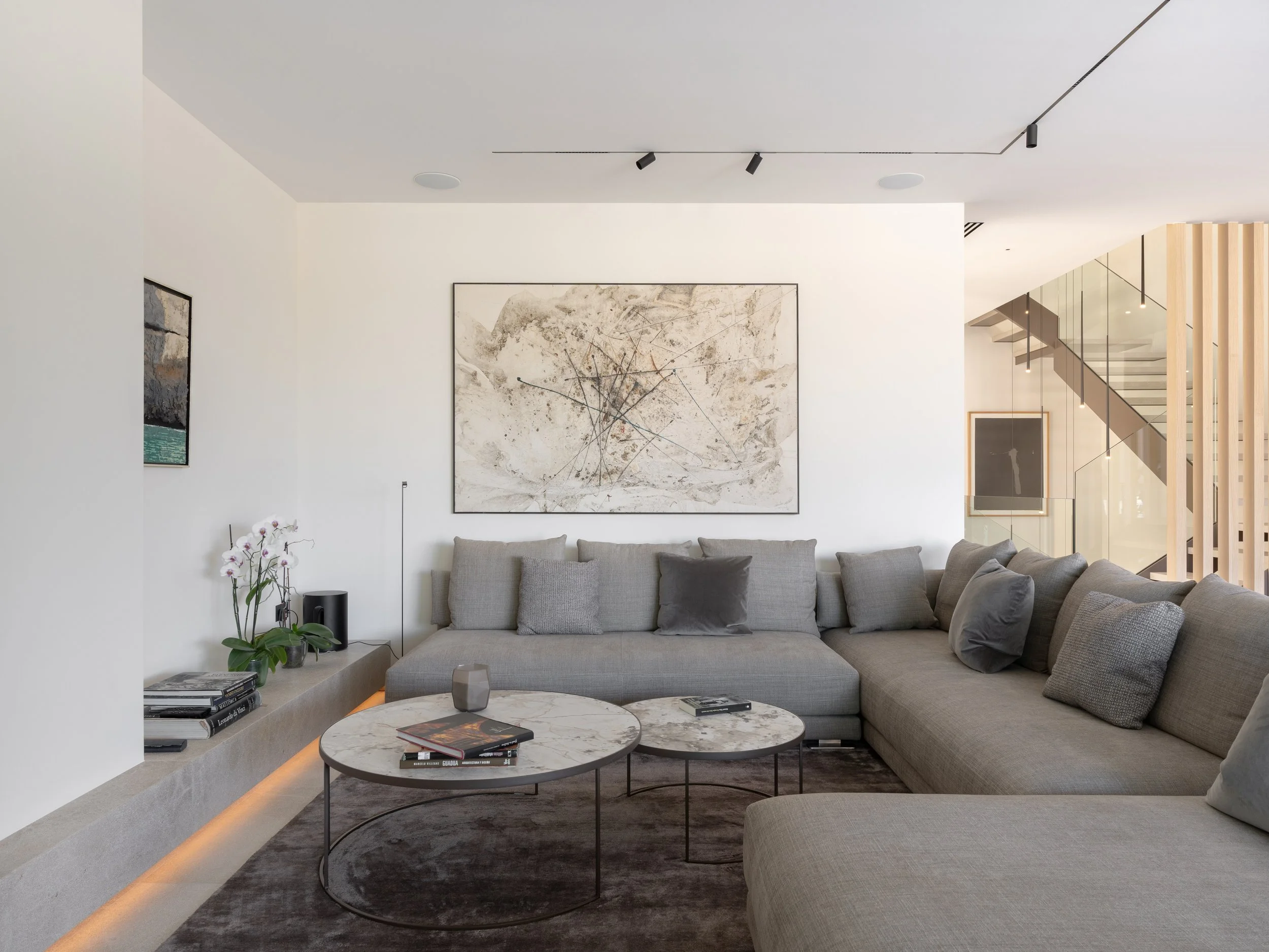 Sala moderna con sofá gris, mesas de centro de mármol, arte abstracto en la pared y escaleras de vidrio a la derecha.