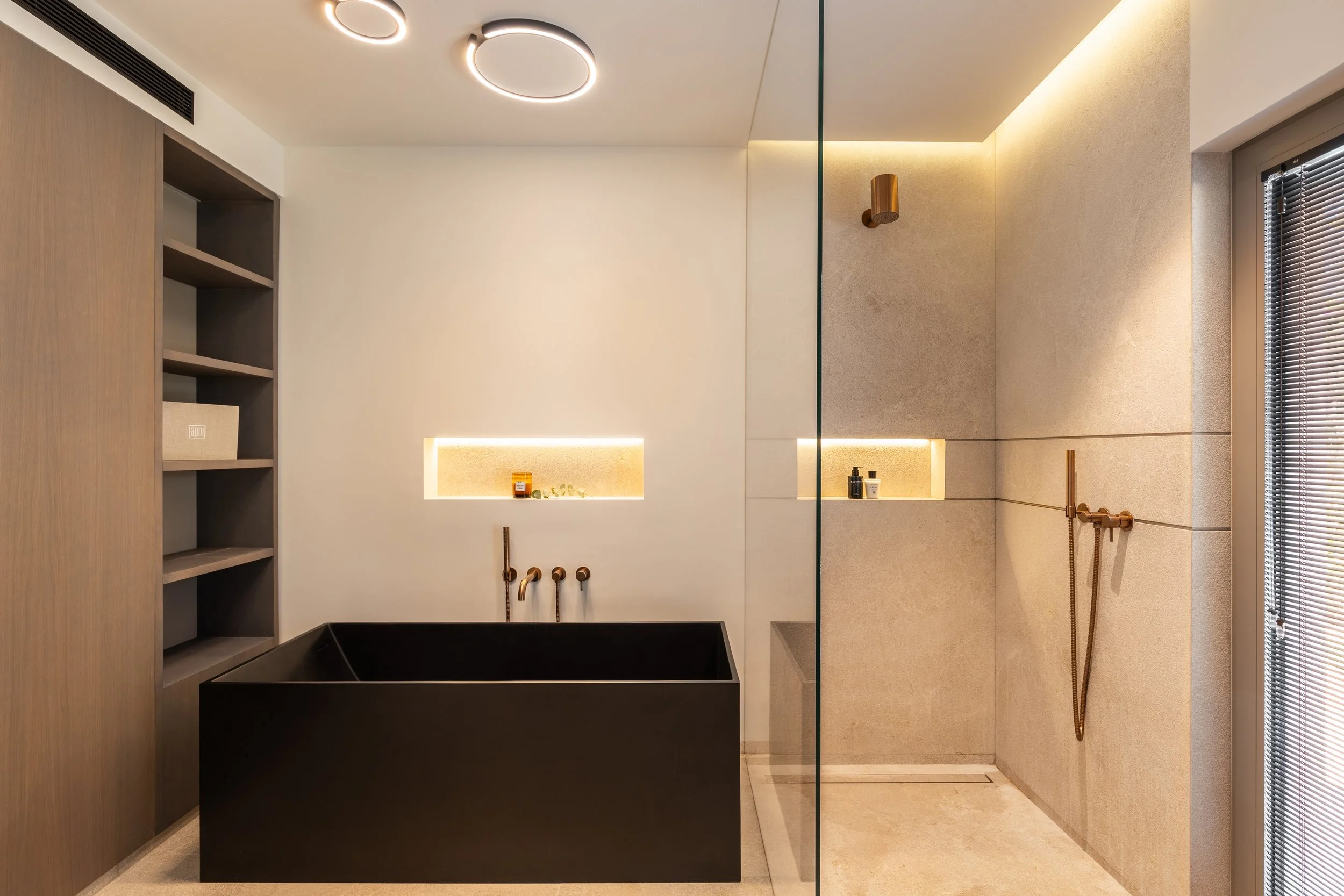Baño moderno con lavabo negro, paredes de azulejos beige y piso de cerámica, estantería de madera y accesorios de cobre, ducha con mampara de vidrio y ventana con cortinas de madera.