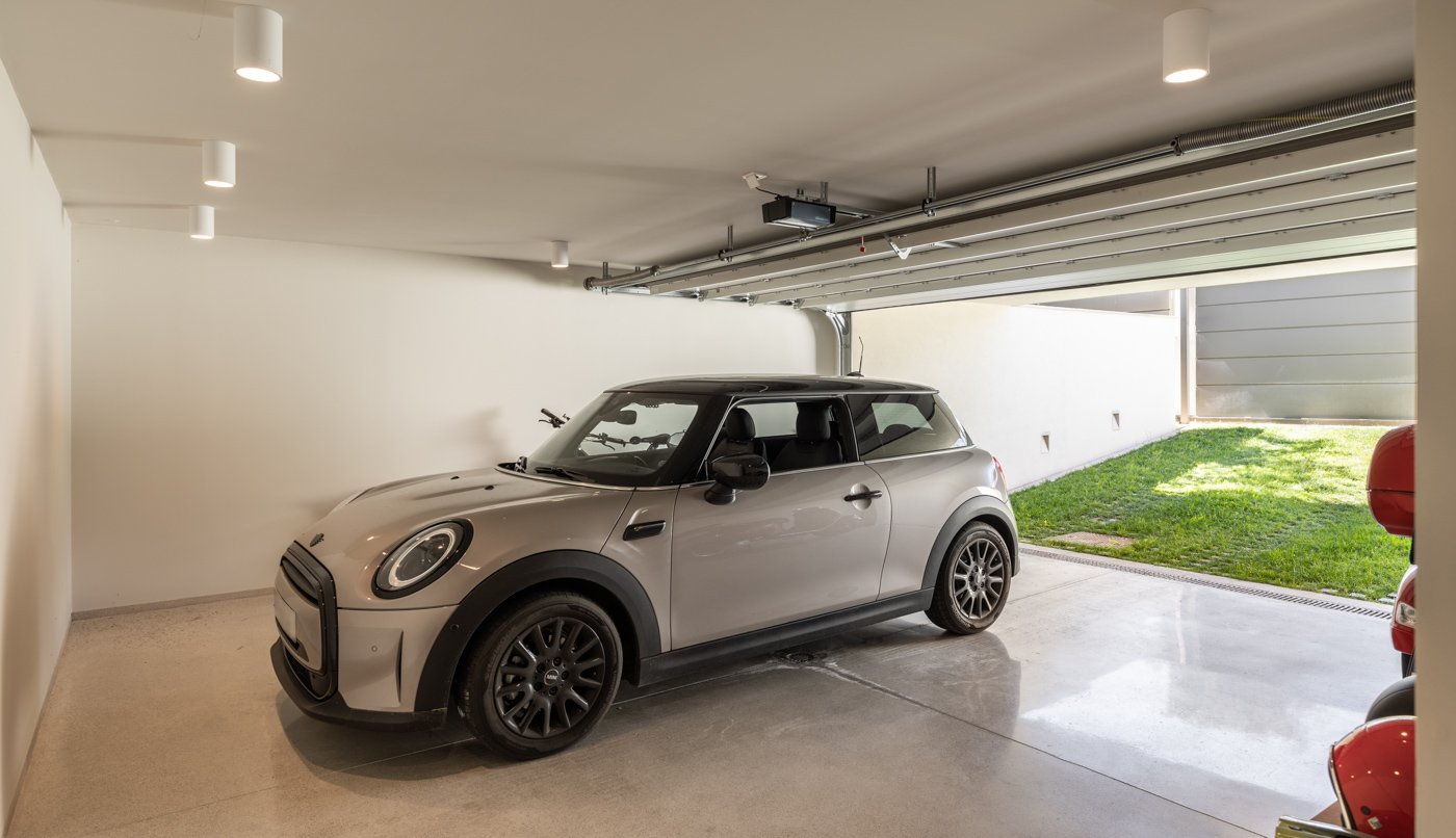 Cochera techada con un auto Mini Cooper de color gris, cerrada y con espacio abierto hacia un jardín exterior con césped.