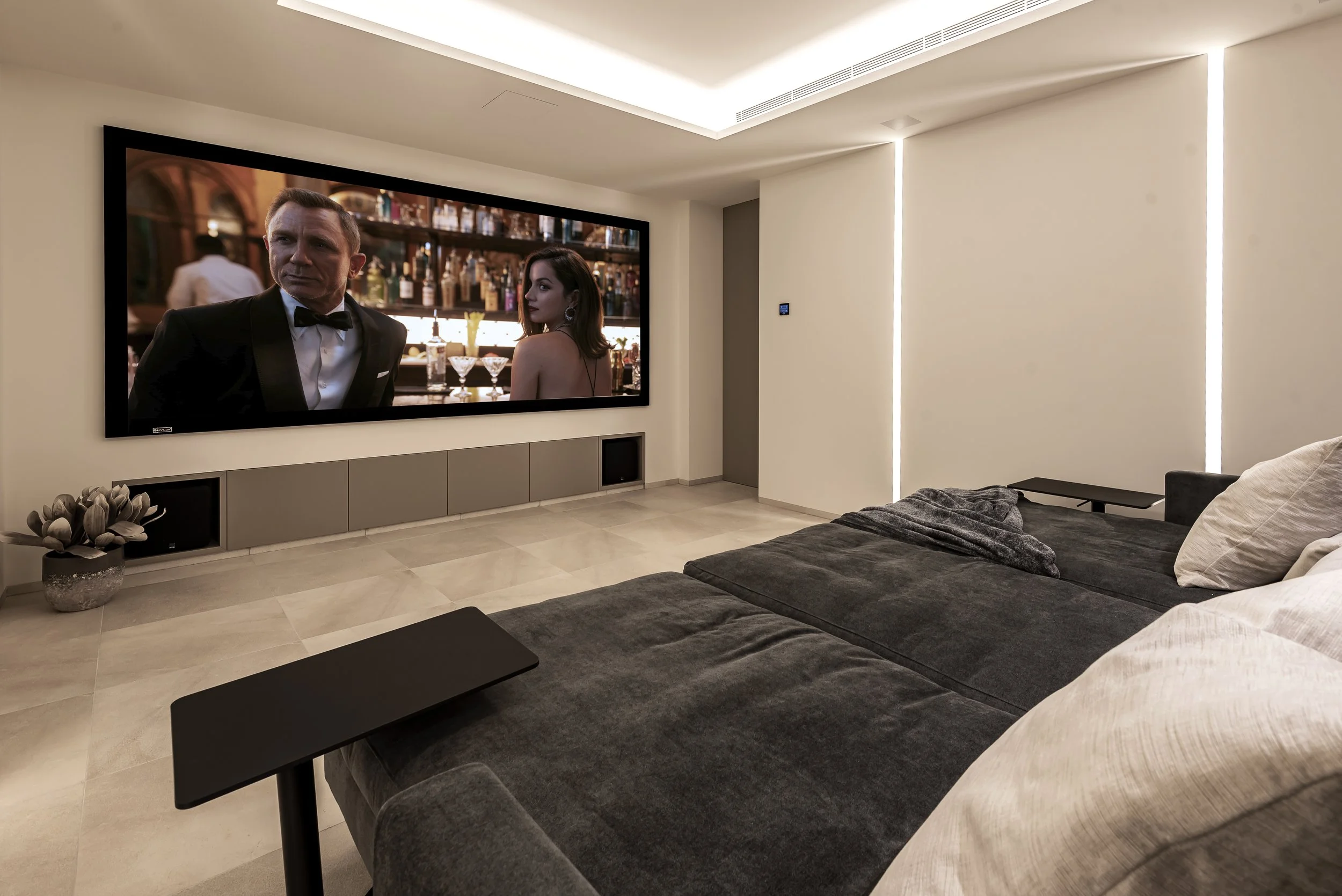 Sala moderna con sofá gris, mesa negra, televisión grande en la pared mostrando una escena de una película o serie con un hombre en un tuxedo y una mujer con vestido de noche, y decoración minimalista en tonos neutros.