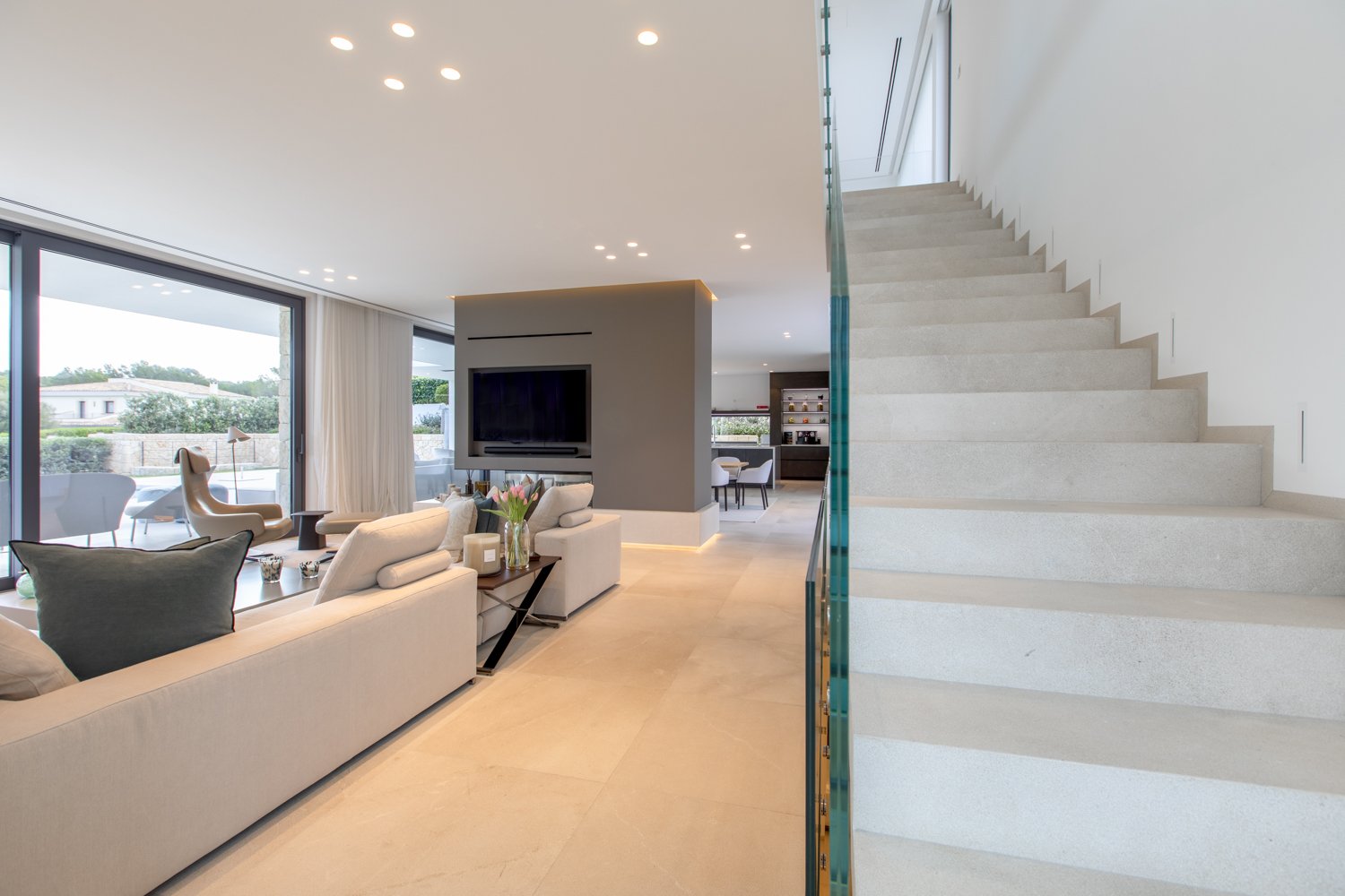 Salón moderno con sofá beige, mesas de centro, televisión en pared, ventana grande con vista al exterior, escaleras de piedra a la derecha, y espacio de comedor en el fondo.