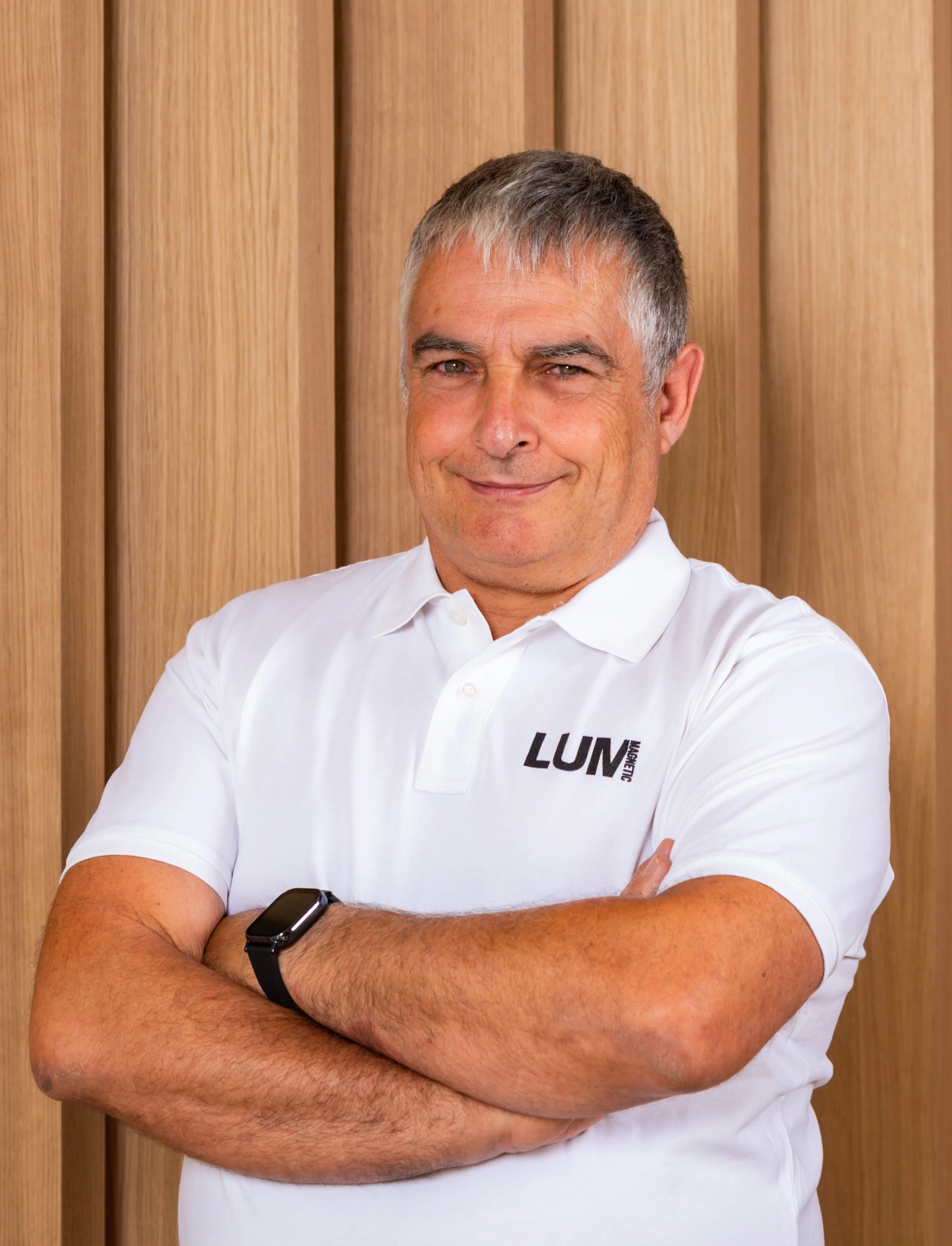 Hombre de cabello rubio y canoso, sonriendo y con los brazos cruzados, usa una camiseta blanca con logotipo y reloj inteligente en la muñeca izquierda, fondo de paneles de madera.