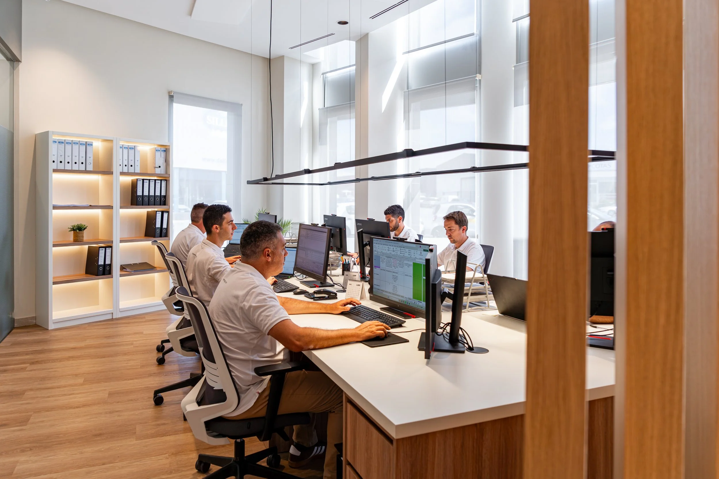 Vista de una oficina moderna con varias personas trabajando en sus computadoras, espacios con grandes ventanas que dejan entrar luz natural y estanterías con carpetas.