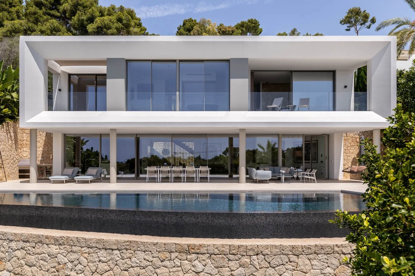 Casa moderna de dos pisos con balcón y vista a una piscina, rodeada de árboles y vegetación.