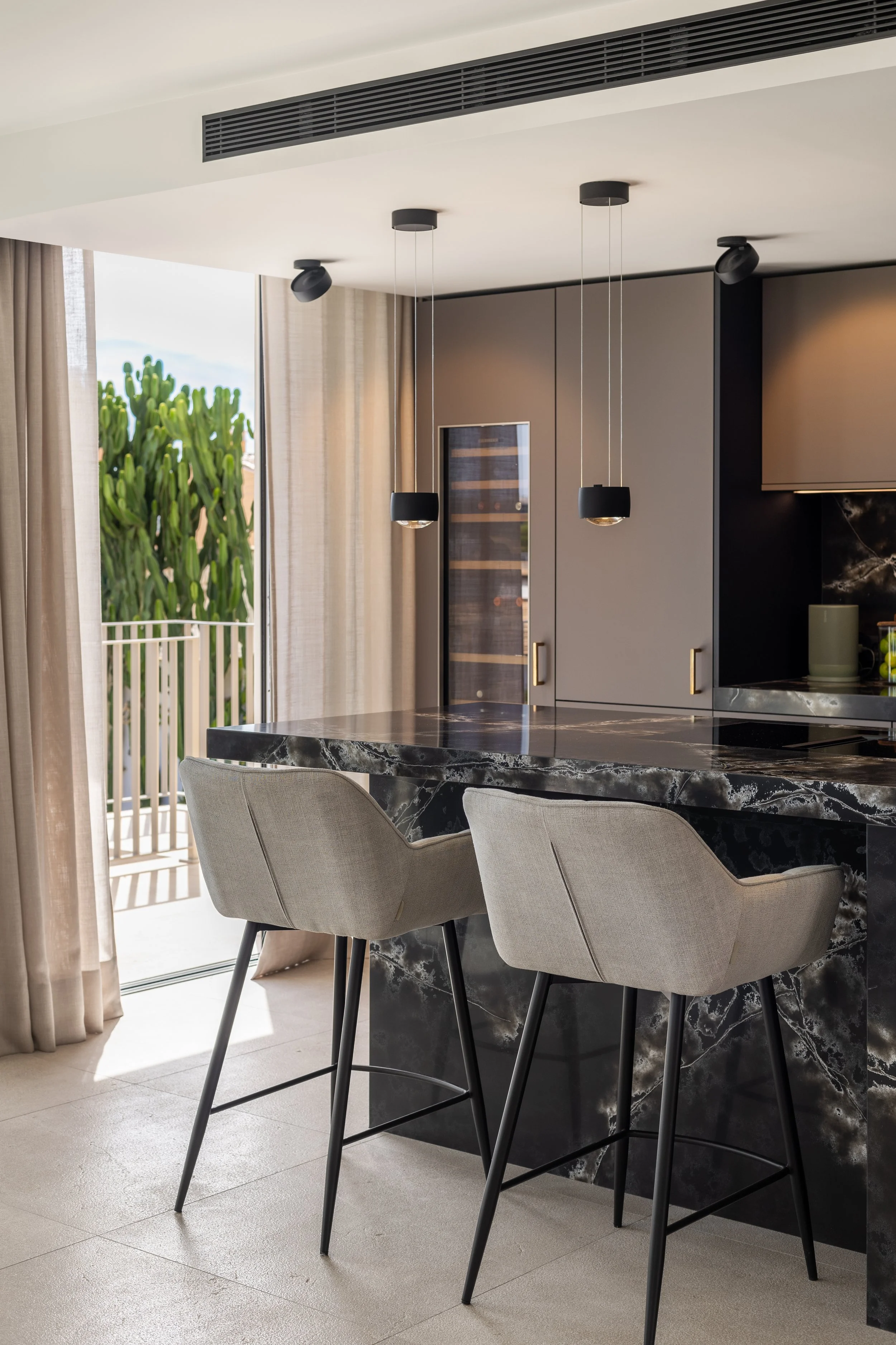 Vista interior de una cocina moderna con una mesa de mármol negro, sillas de color gris claro, armarios grises, y una puerta que lleva a un balcón con plantas verdes y cortinas beige, iluminación moderna y ventilación en el techo.