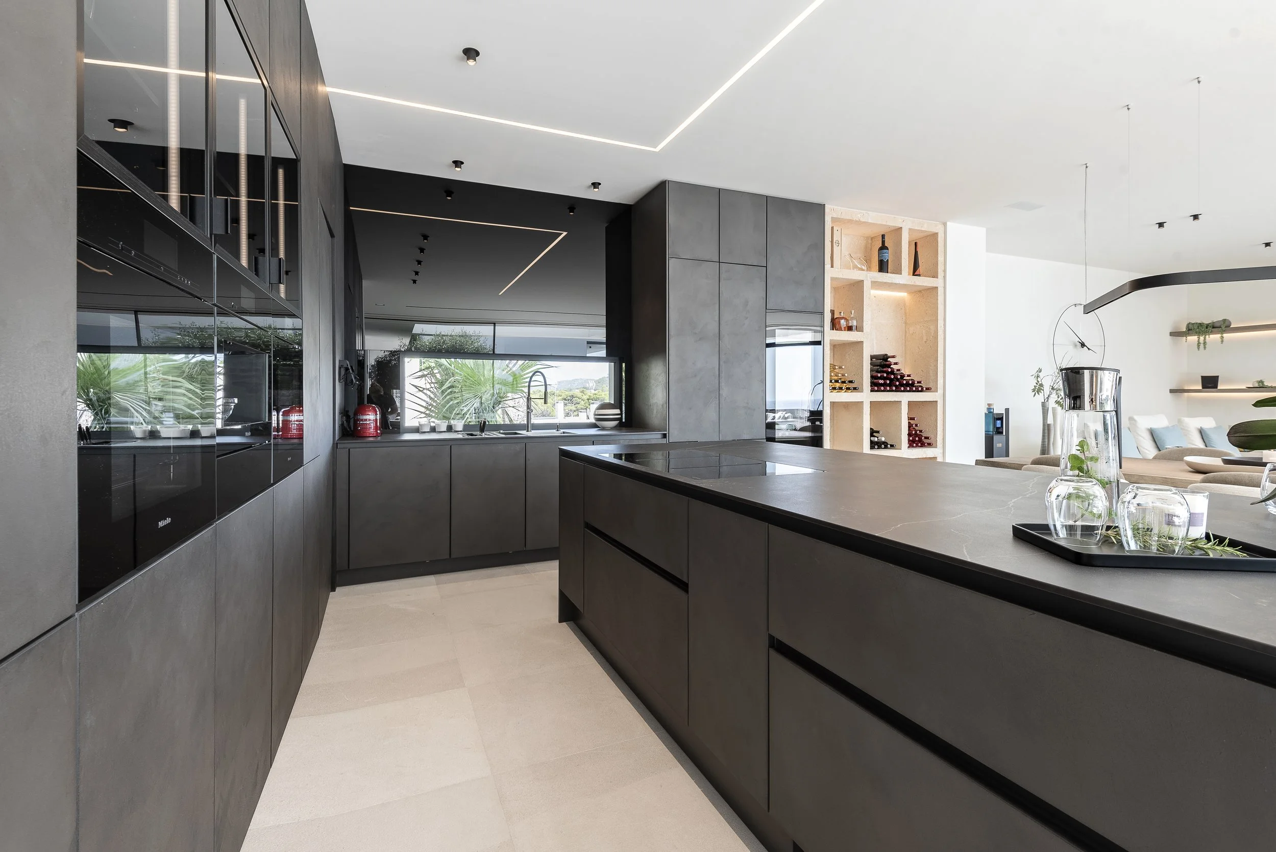 Cocina moderna con armarios de color gris oscuro, encimera de piedra y estanterías con vinos y decoraciones, área de comedor con mesas y sillas, iluminación moderna en el techo, ventanas grandes con vista a áreas verdes.
