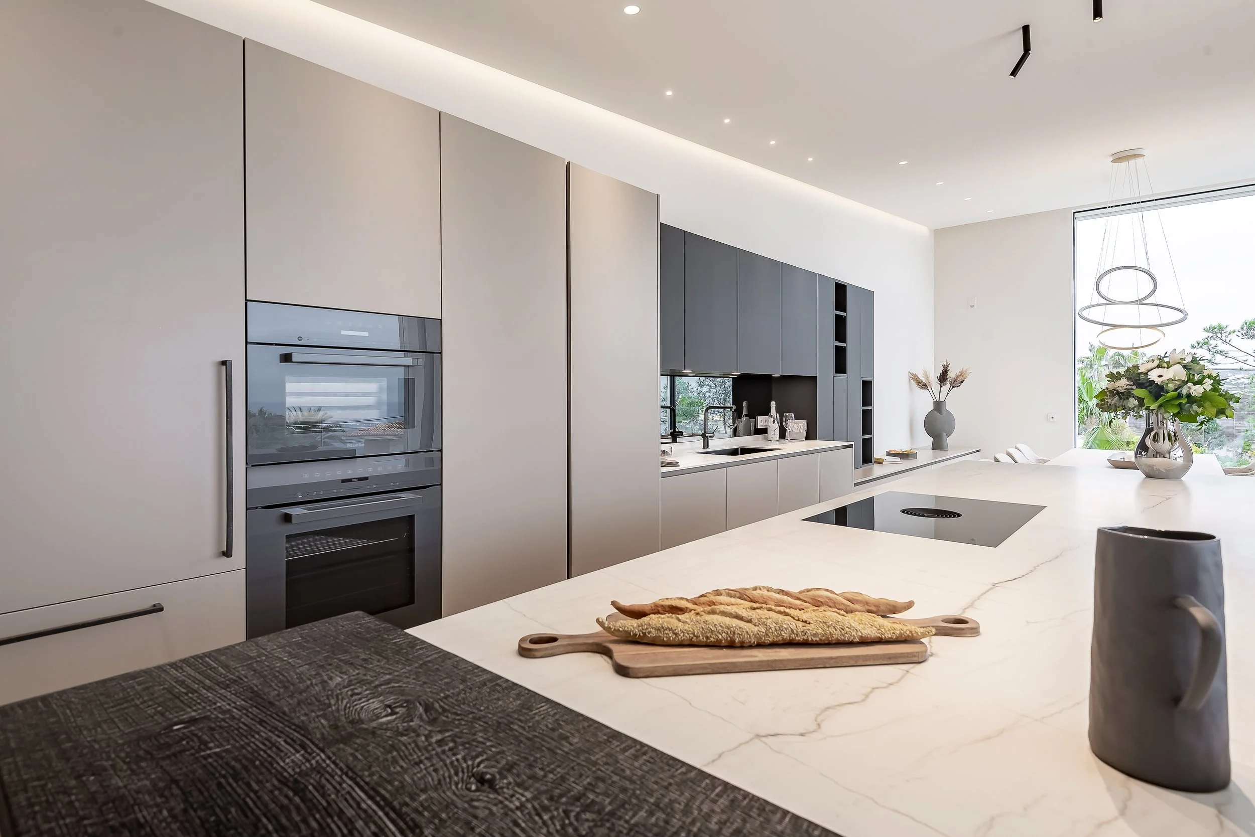 Cocina moderna con lavavajillas, horno, gabinetes grises, ventana grande, mesa de marfil con florero y utensilios para cocinar.