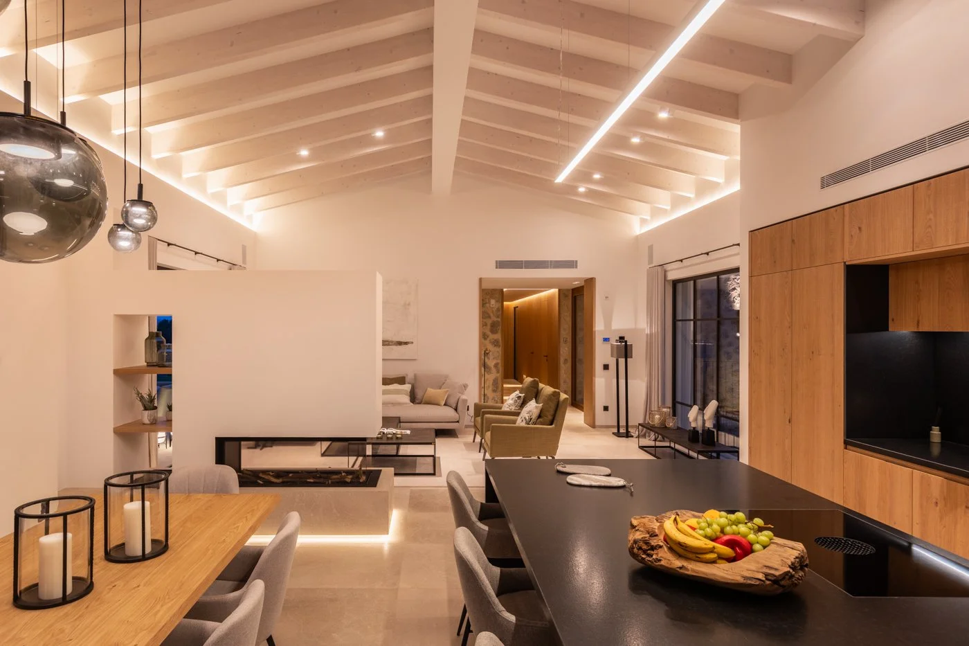 Sala de estar y comedor en una casa moderna con techo de vigas blancas, mobiliario en tonos neutros, y una cocina con isla de piedra negra y madera natural, decorada con frutas en una bandeja en la encimera.