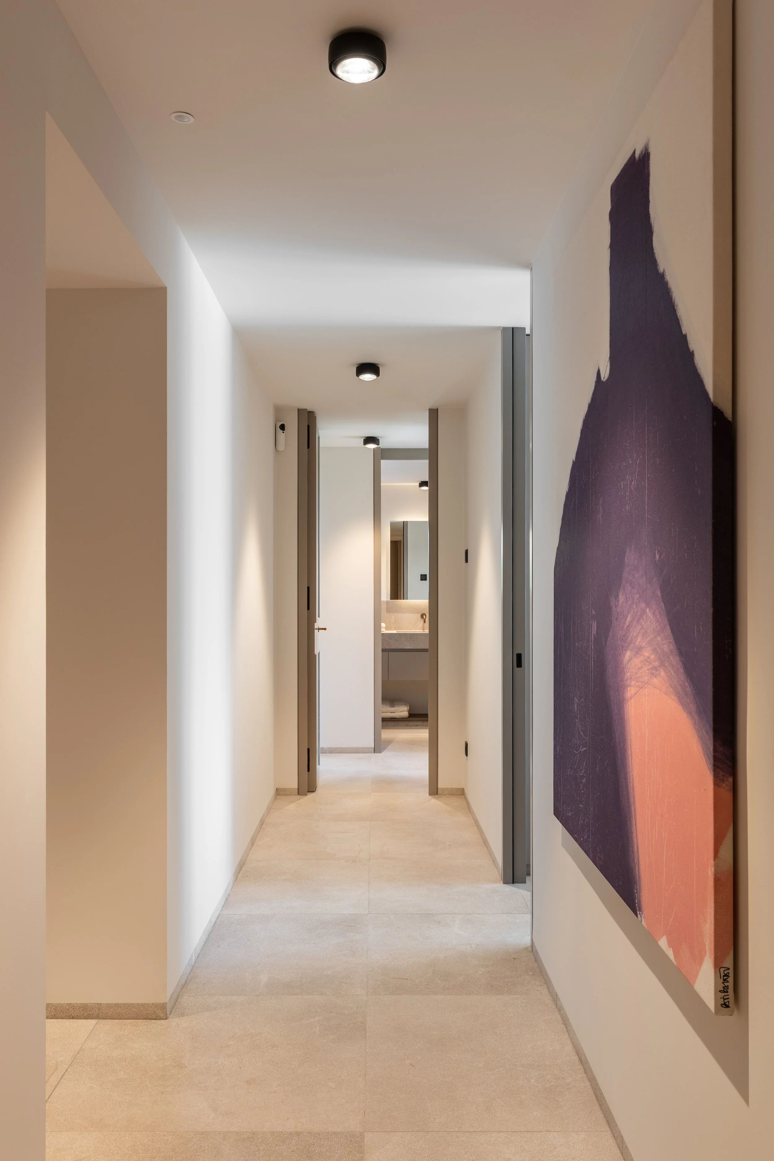 Pasillo interior con paredes blancas, iluminación en el techo, y una pintura grande de montañas en la pared derecha.