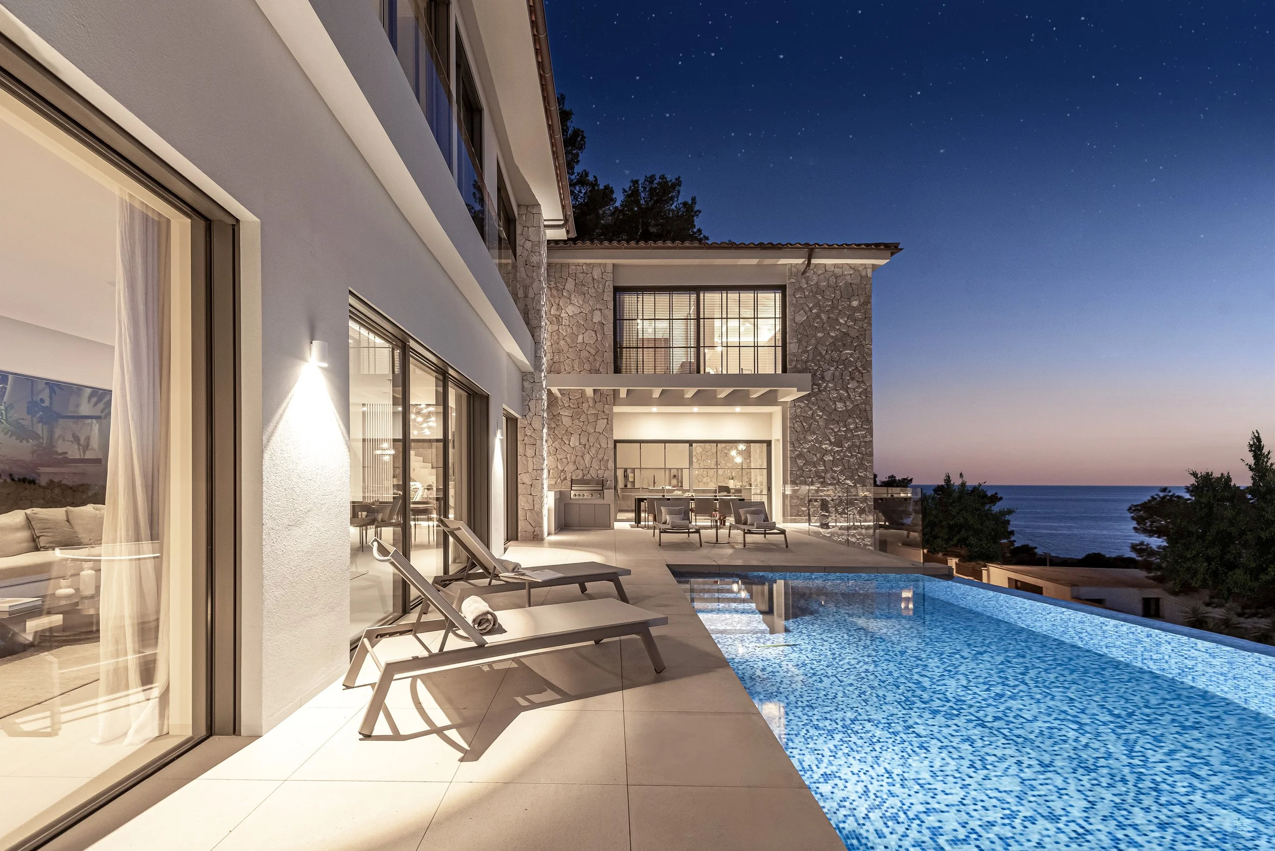 Vista de una casa moderna con piscina y terraza, con vista al mar durante la noche, iluminada con luz cálida