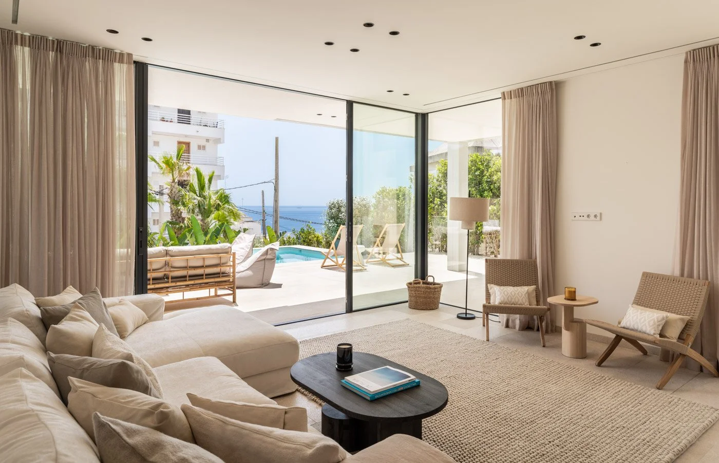 Interior de sala moderna con sofá beige, mesa de centro negra, sillas de madera, alfombra de fibras naturales, cortinas beige, vista a una terraza y piscina con plantas y vista al mar.