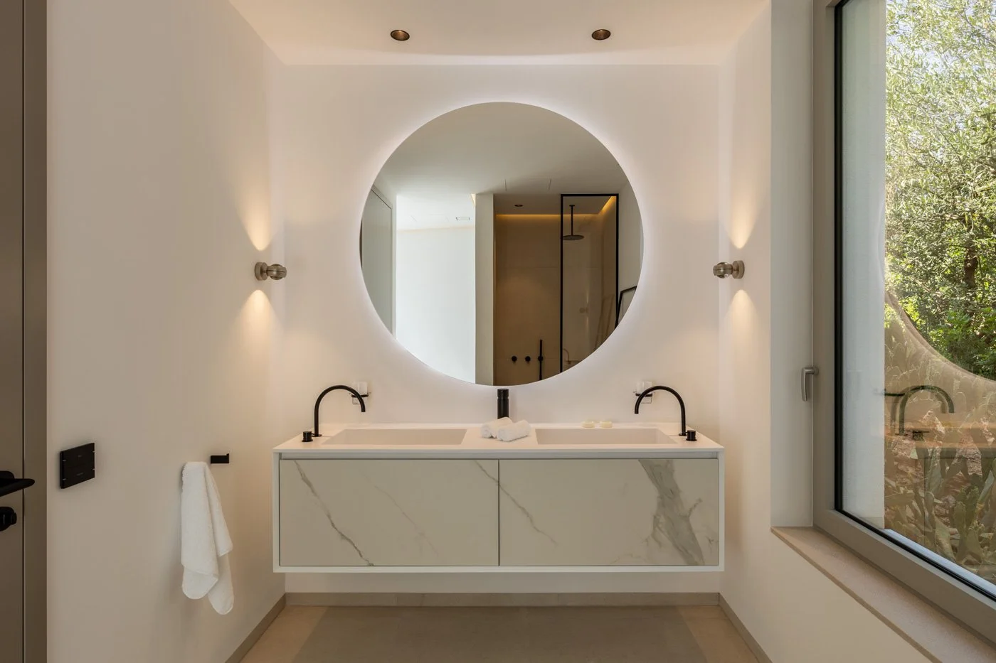 Lavabo doble con encimera de mármol, espejo grande redondo, ventanas grandes con vista a árboles, paredes blancas, luces empotradas en techo y paredes, toallas blancas