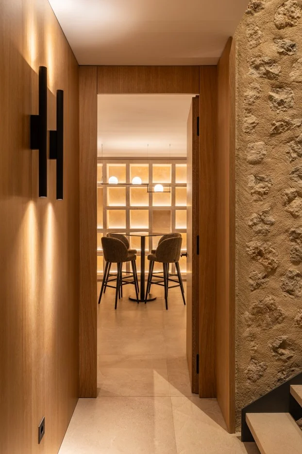Vista desde una entrada de puertas de madera hacia una sala con sillones y una mesa en el centro, fondo con pared de bloques de piedra y lámparas redondas que iluminan suavemente.